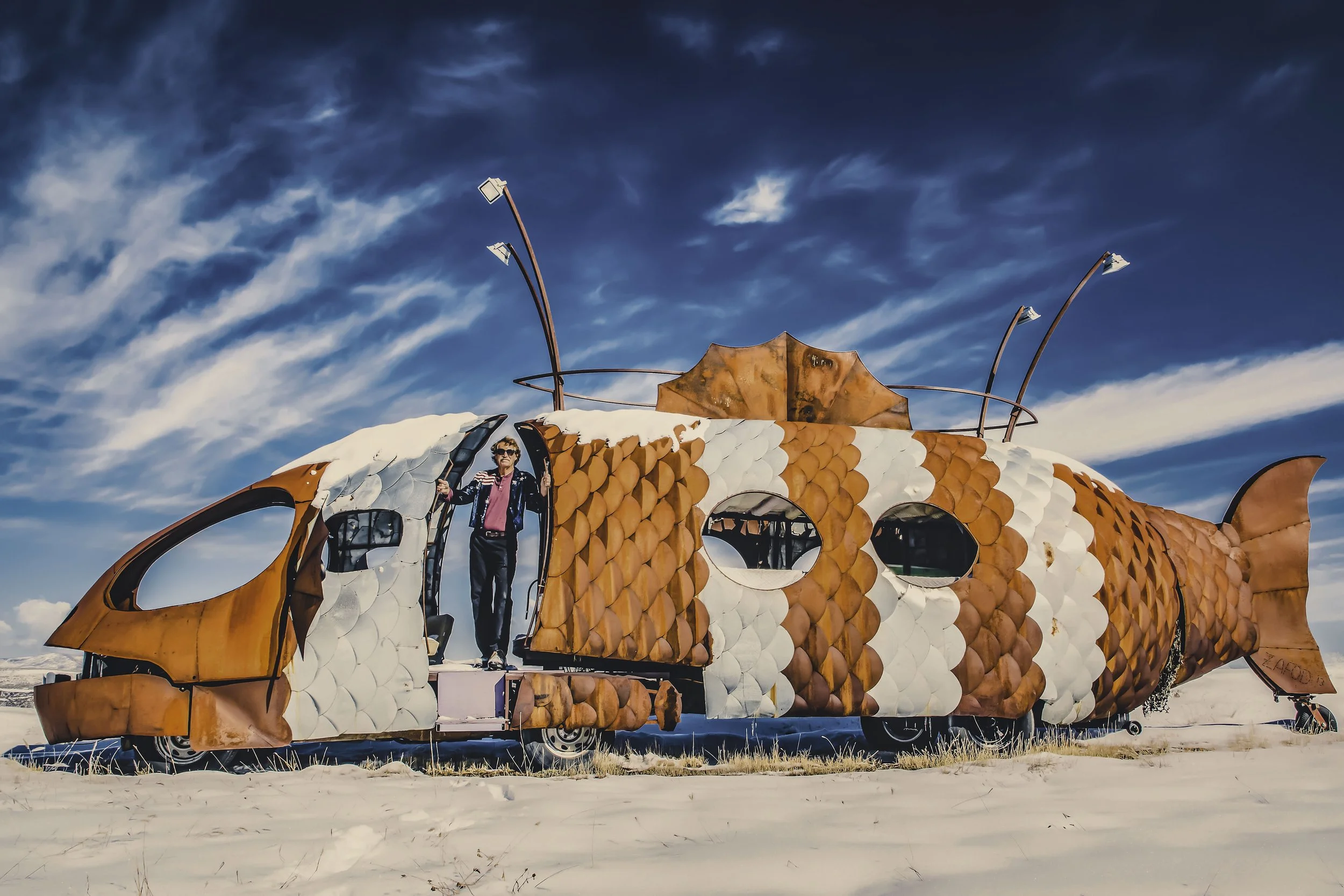 A person standing inside a large, animal-shaped sculpture resembling a fish or dragon, made of orange and white panels, with a clear sky and clouds in the background.