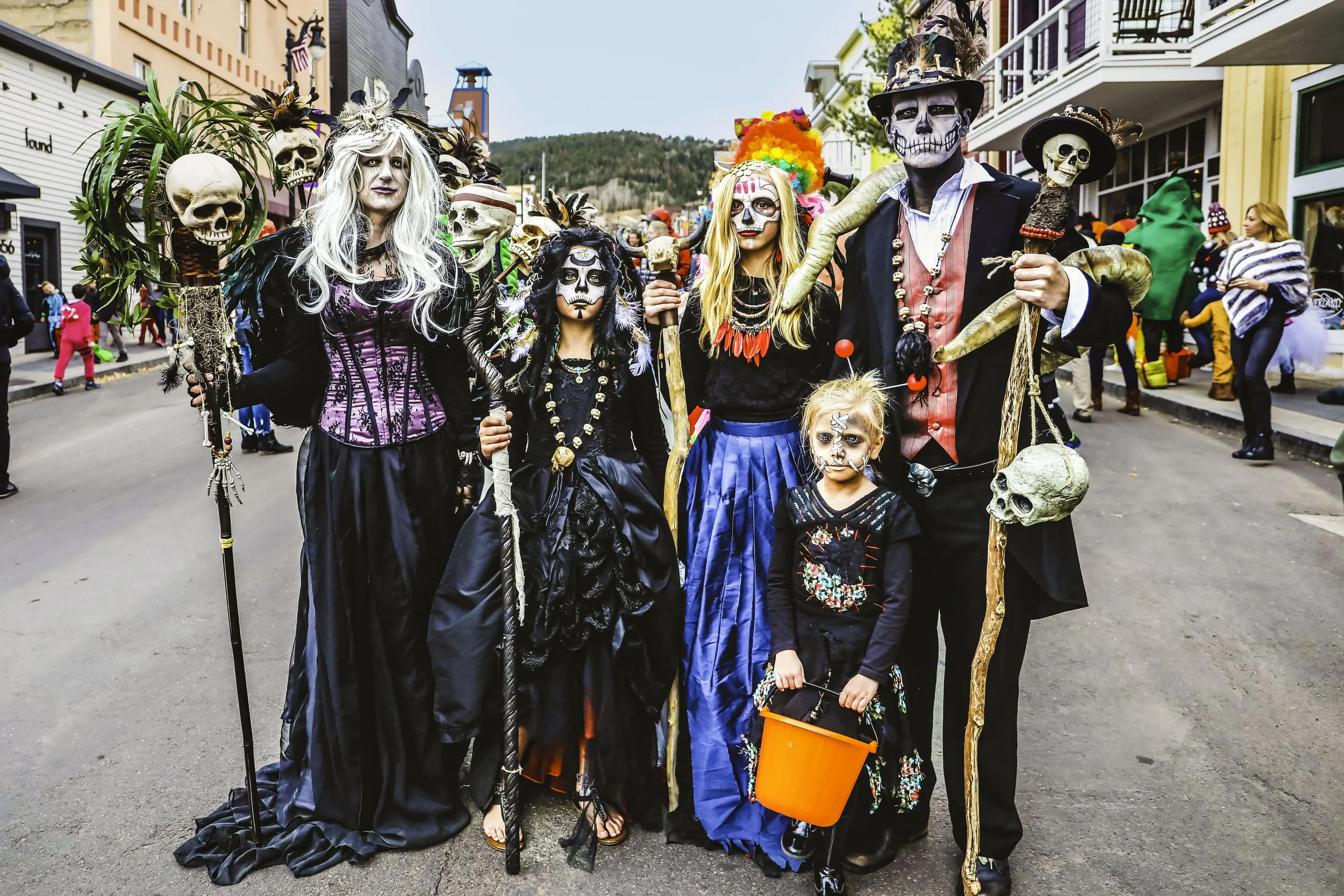 Group of adults and children in Halloween costumes and makeup, including skeletons and Day of the Dead characters, standing on a city street during a Halloween parade.
