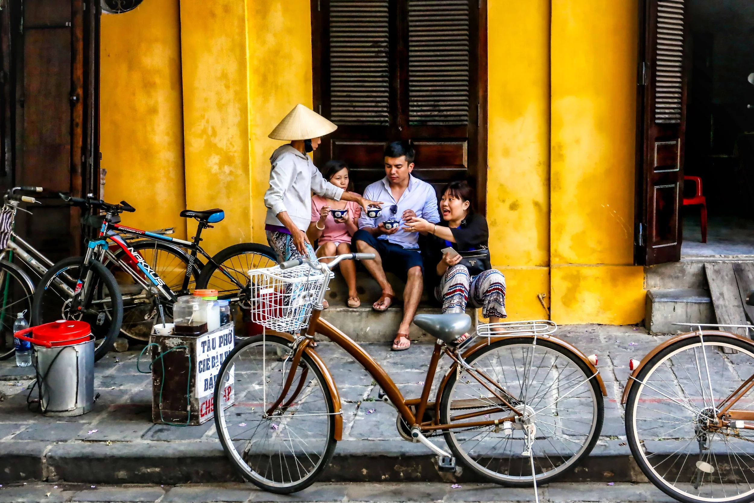 Four people sitting outside a yellow building, engaging in conversation, with bicycles in the foreground. One woman is wearing a traditional conical hat and addressing the others, who are holding bowls. Bicycles and some containers are visible on the