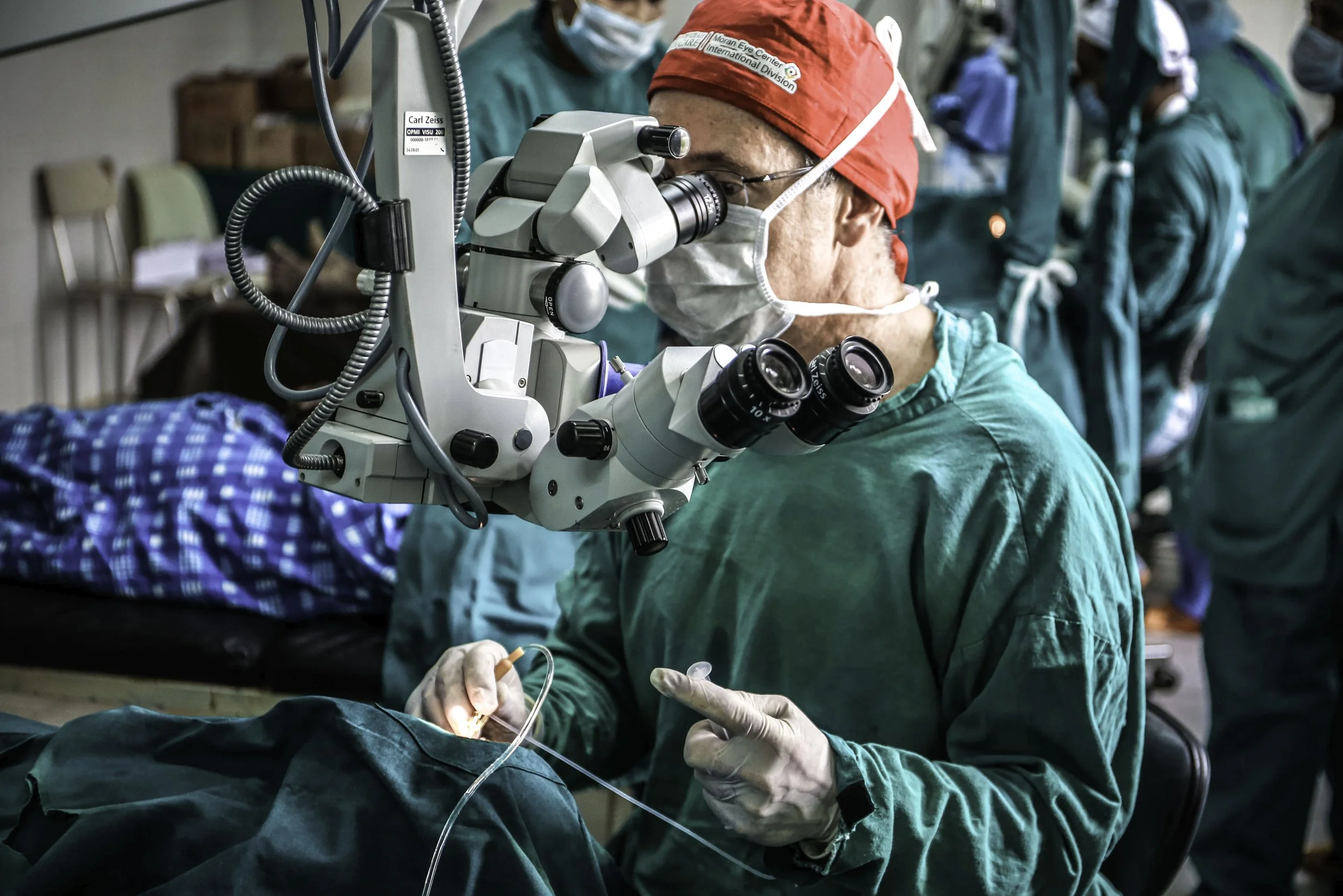 A medical professional wearing green surgical scrubs, a red surgical cap, and a face mask is using a microscope during surgery. There are other medical staff in similar attire in the background.