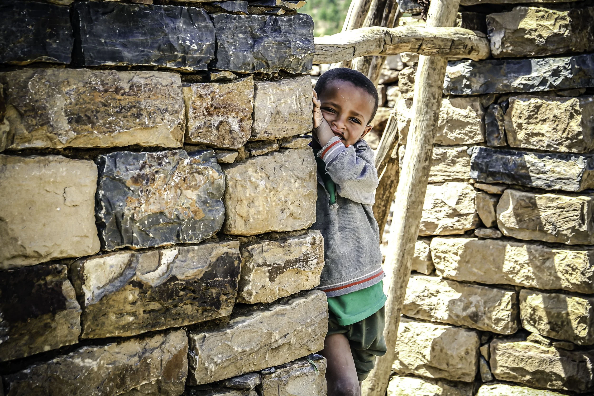 A young boy wearing a gray hoodie and shorts peeks through a gap in a stone wall, smiling at the camera.