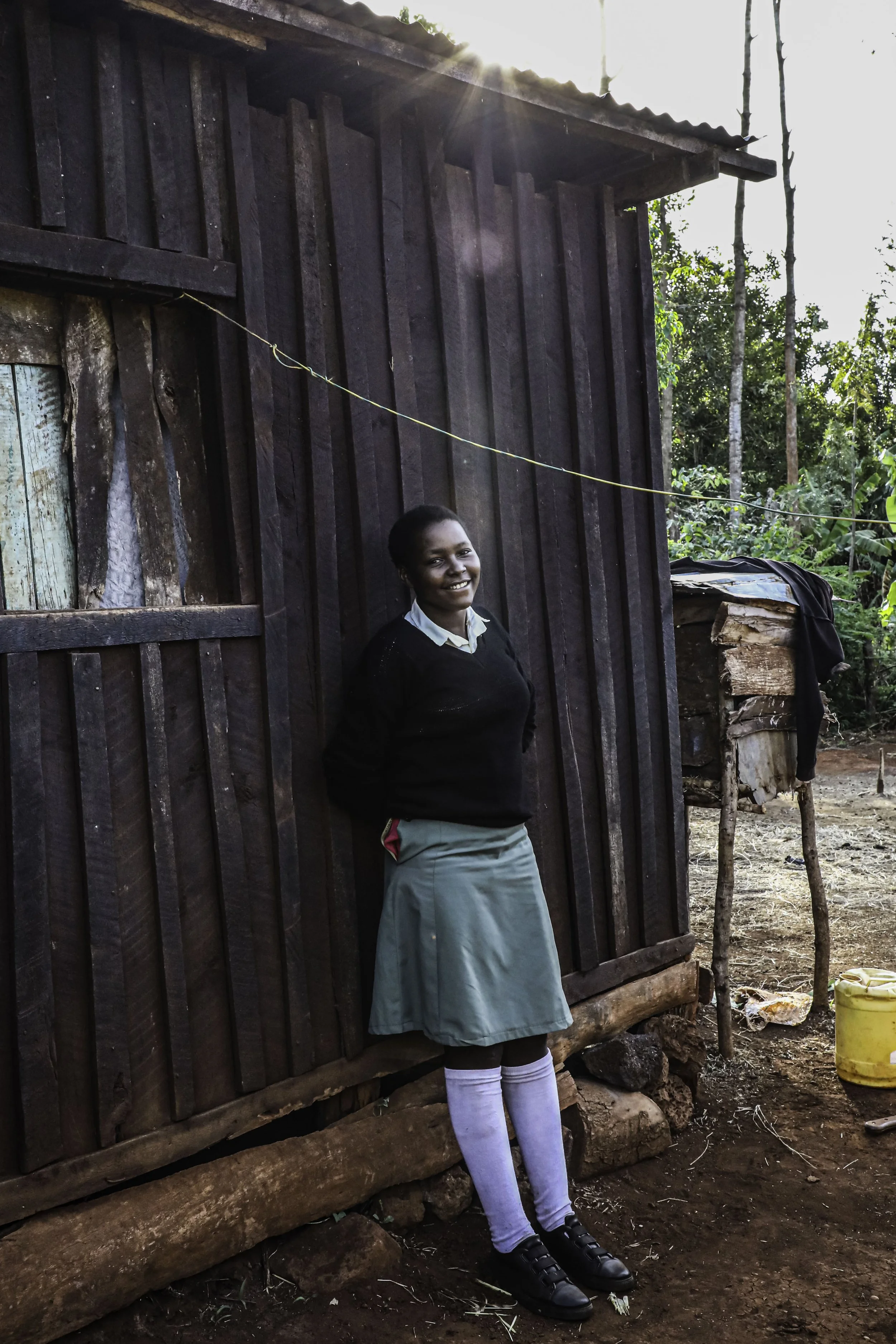 A young girl in a school uniform standing outside next to a dark wooden building, smiling at the camera. The girl is wearing a light blue skirt, white socks, and black shoes. The background shows a rural setting with trees and a yellow container on t