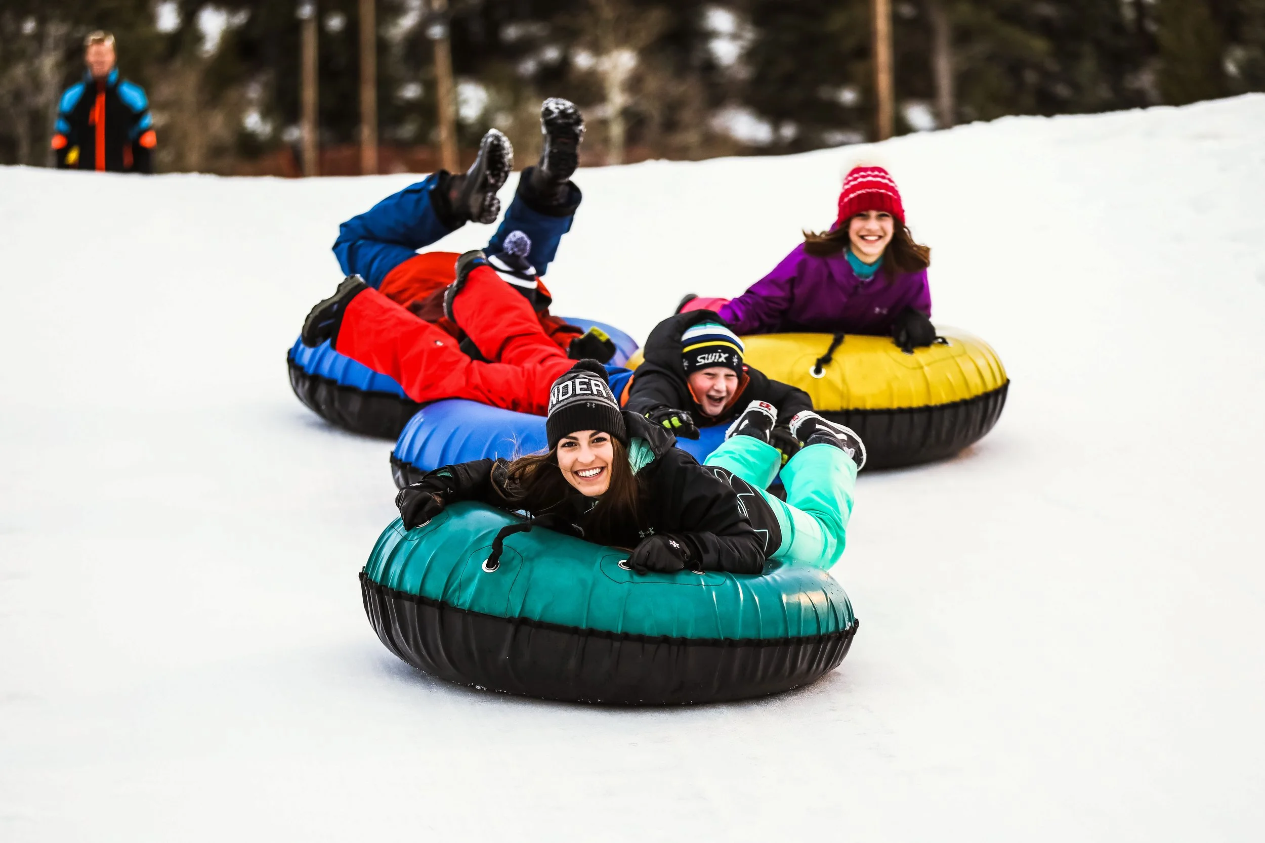 Four children with winter clothing enjoying sledding down a snowy hill on inflatable tubes, with a woman standing in the background.