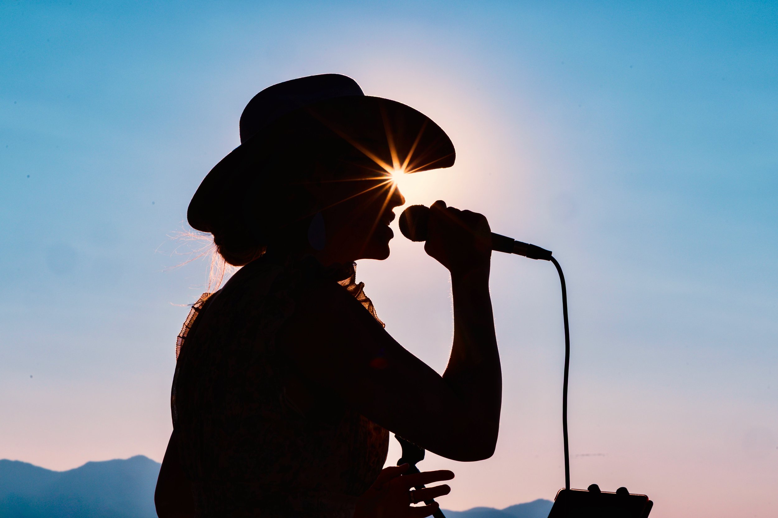 Silhouette of a woman wearing a hat, singing into a microphone with sun flare behind her, outdoors during sunset or sunrise, mountains in the background.