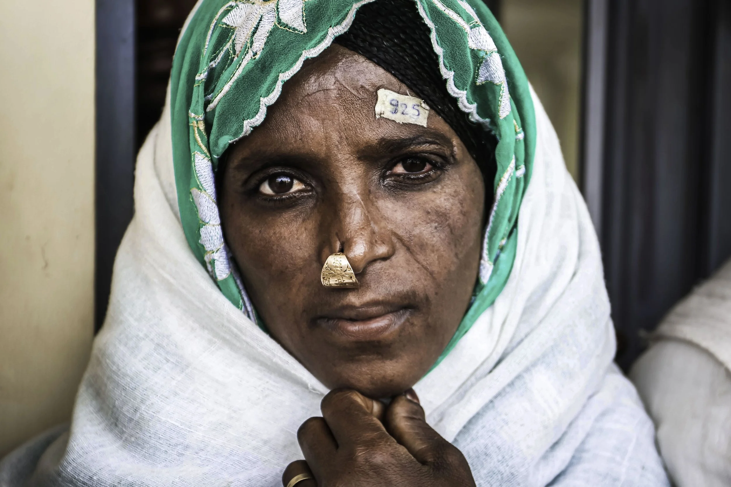 Close-up of a woman wearing a green and white headscarf with embroidered details, a black head covering underneath, and gold jewelry, looking directly at the camera with her hand near her chin.
