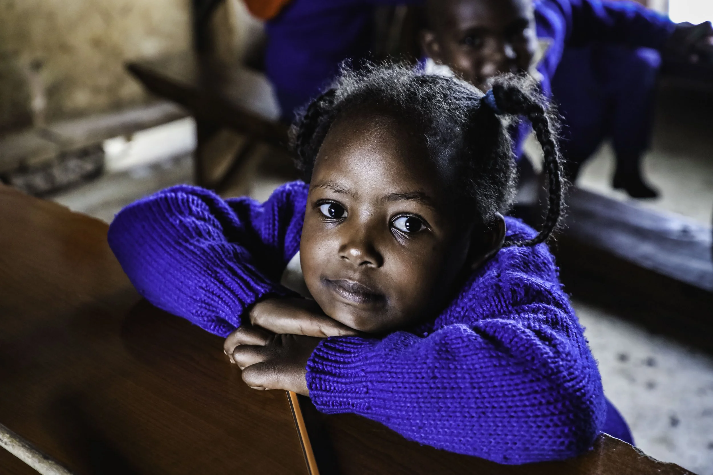 A young girl with curly hair and bright eyes wearing a blue sweater, resting her head on her arms on a wooden desk, with another girl in the background smiling.