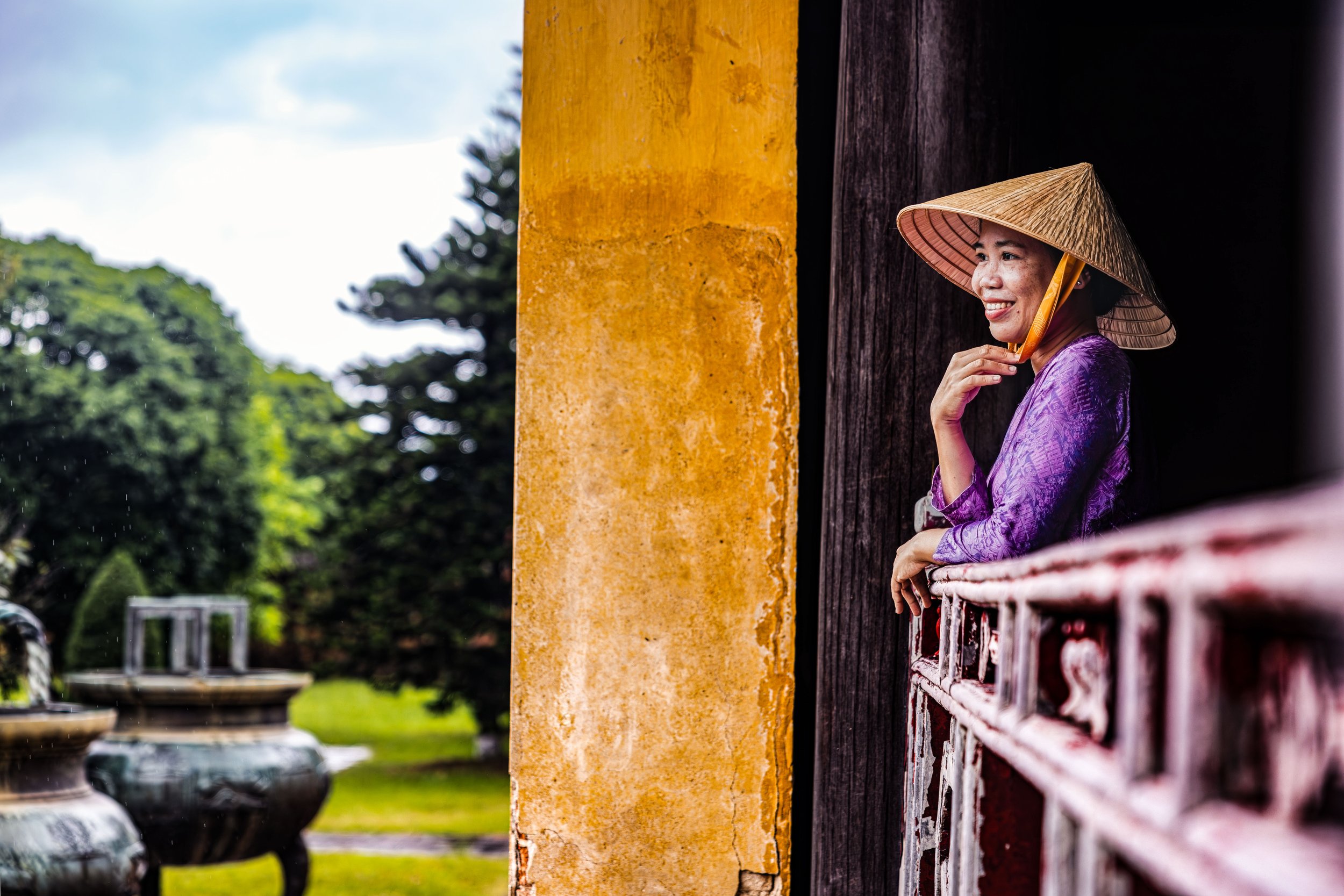 A woman wearing a traditional Asian conical hat and purple attire sitting on a balcony with a wooden railing, smiling and looking out over a garden with trees and decorative pots.