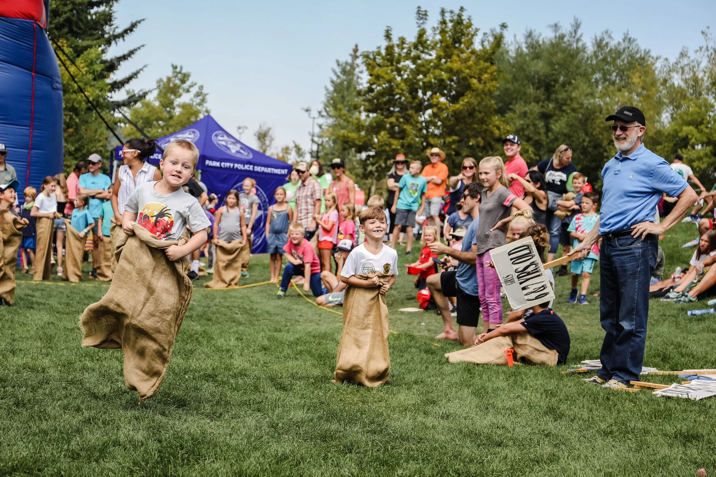 Children participate in a sack race at an outdoor event, surrounded by a crowd of spectators and a park police tent, on a grassy field with trees in the background.