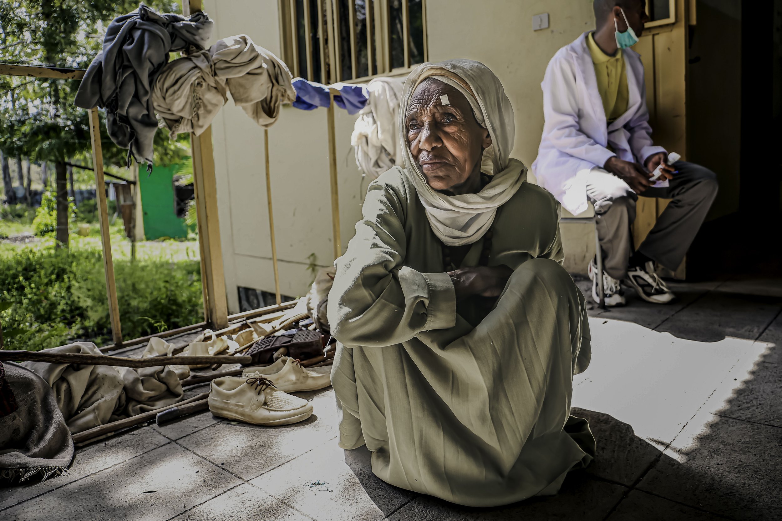 An elderly woman wearing a headscarf sits outdoors on a tiled floor, surrounded by scattered shoes and personal belongings. In the background, a younger man wearing a face mask and white jacket is seated against the wall, holding a small object.