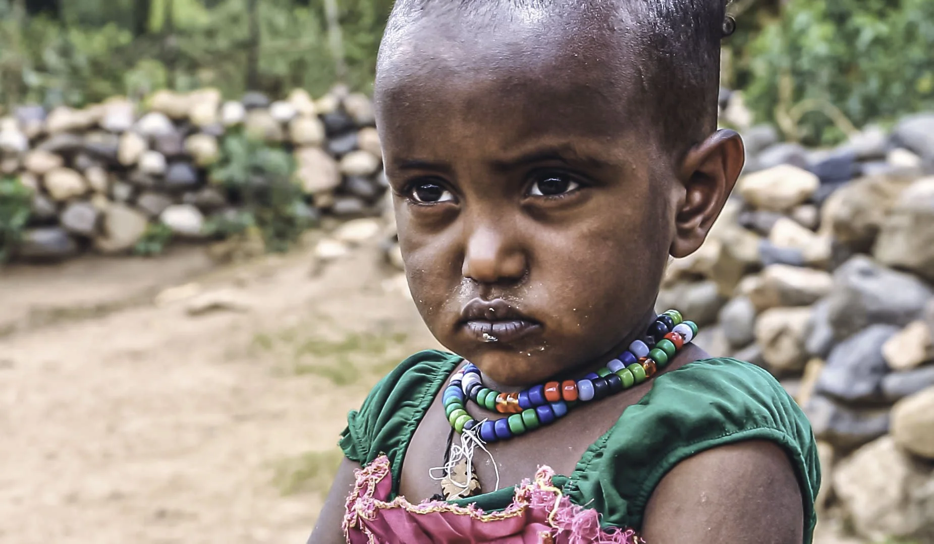 A young child with dark skin and large, expressive eyes, wearing colorful beaded necklaces and a green and pink dress outdoors in a rural setting with a stone wall and trees in the background.