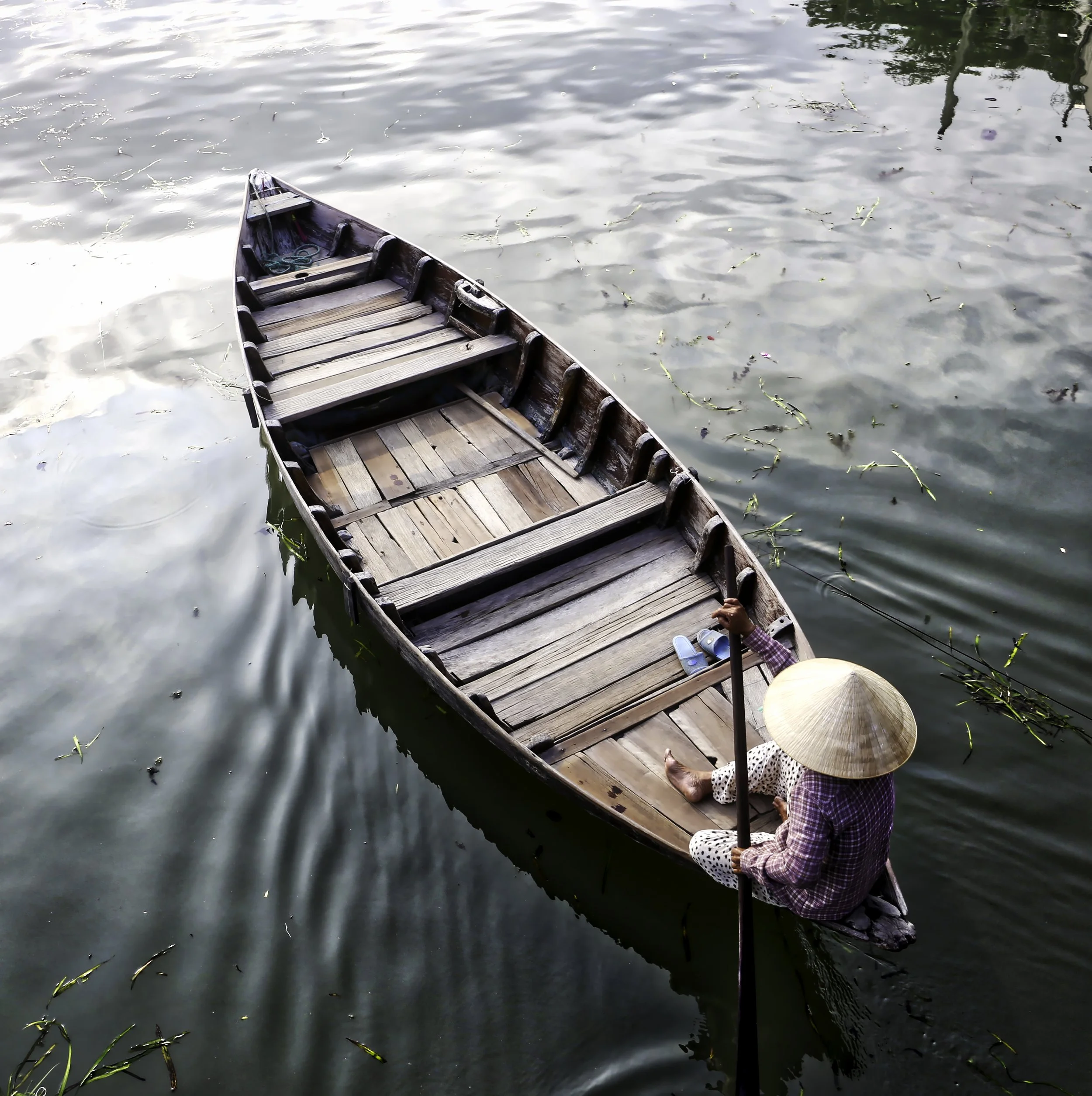 A person wearing a wide-brimmed hat and polka dot clothing sitting in a small wooden boat on a body of water, with greenery and trees reflected in the water and some floating debris or plants.