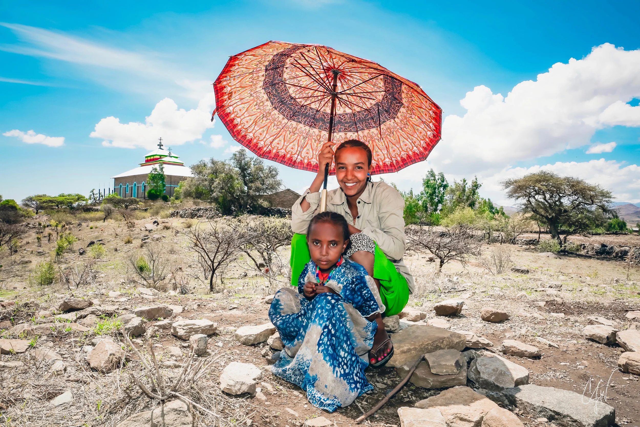 Two young girls sitting outdoors on rocky ground, with one holding a large decorative umbrella. A church or similar building is visible in the background among trees, under a bright blue sky with clouds.