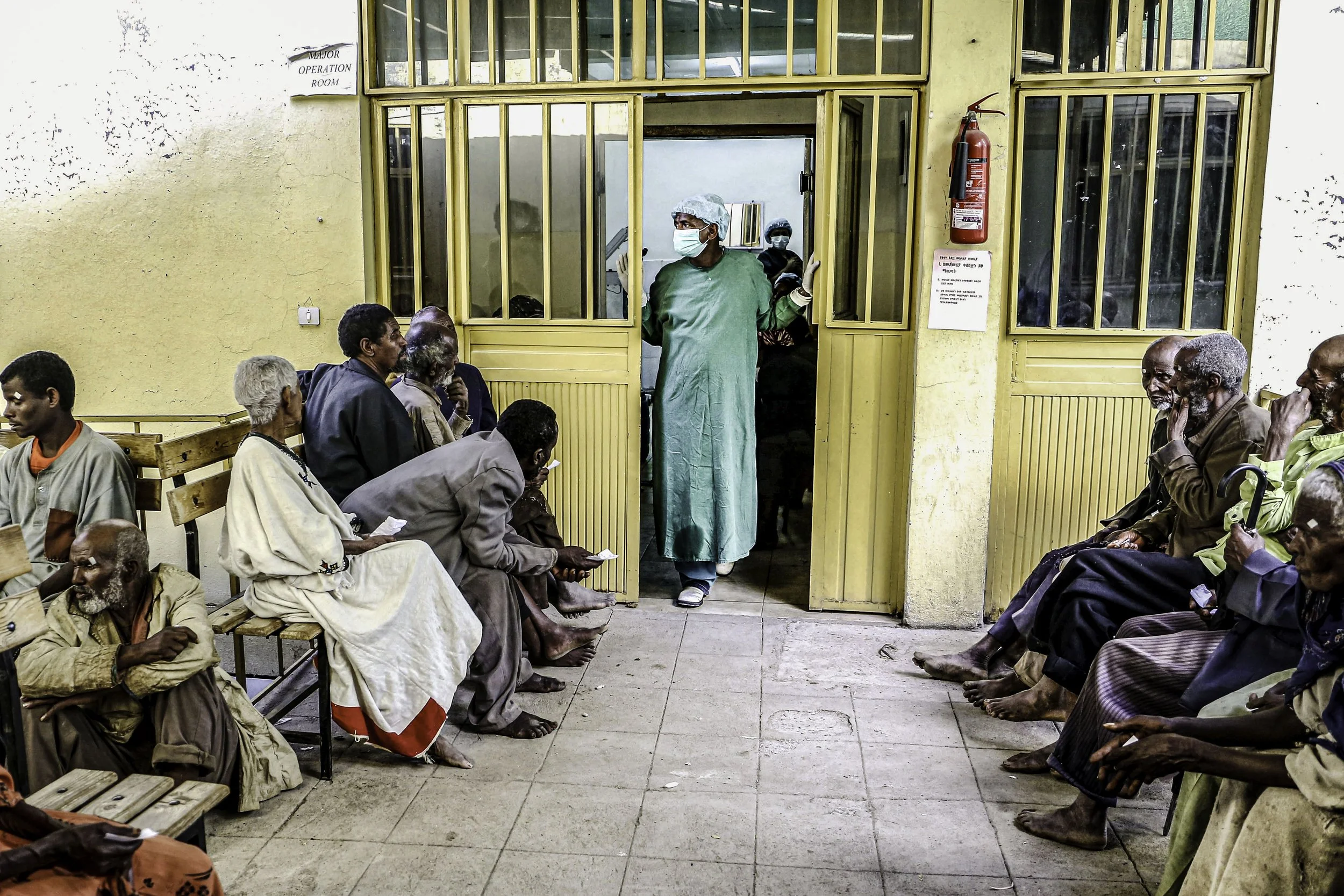 A group of people sitting on benches inside a hospital waiting area, some with bandages and others with visible injuries. A healthcare worker in scrubs, mask, and gloves stands in the doorway, gesturing with hands. The room has yellow walls, wooden b