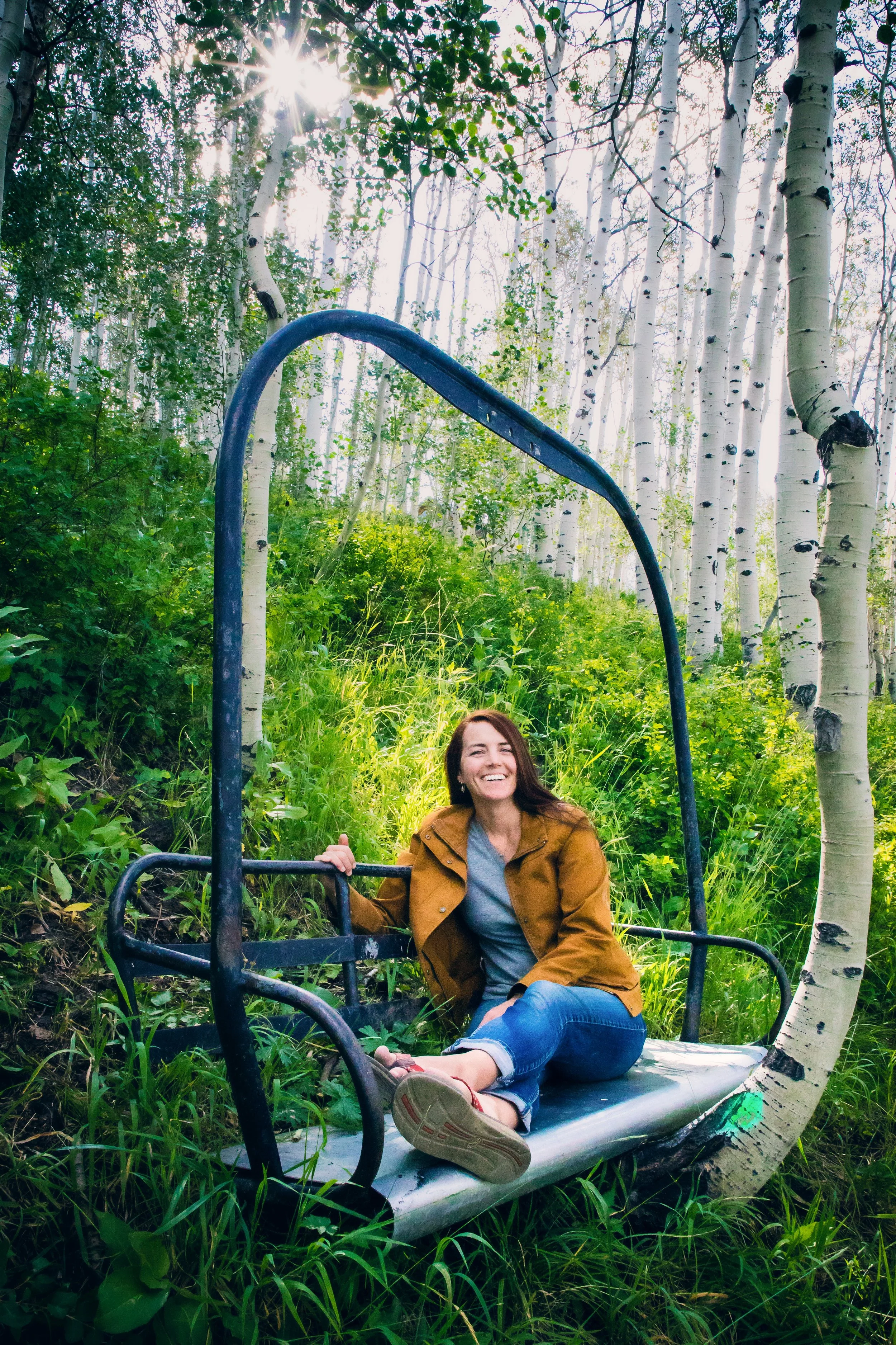 Smiling woman sitting on a metal slide in a forest, surrounded by tall white-barked trees and green foliage, with sunlight filtering through the branches.