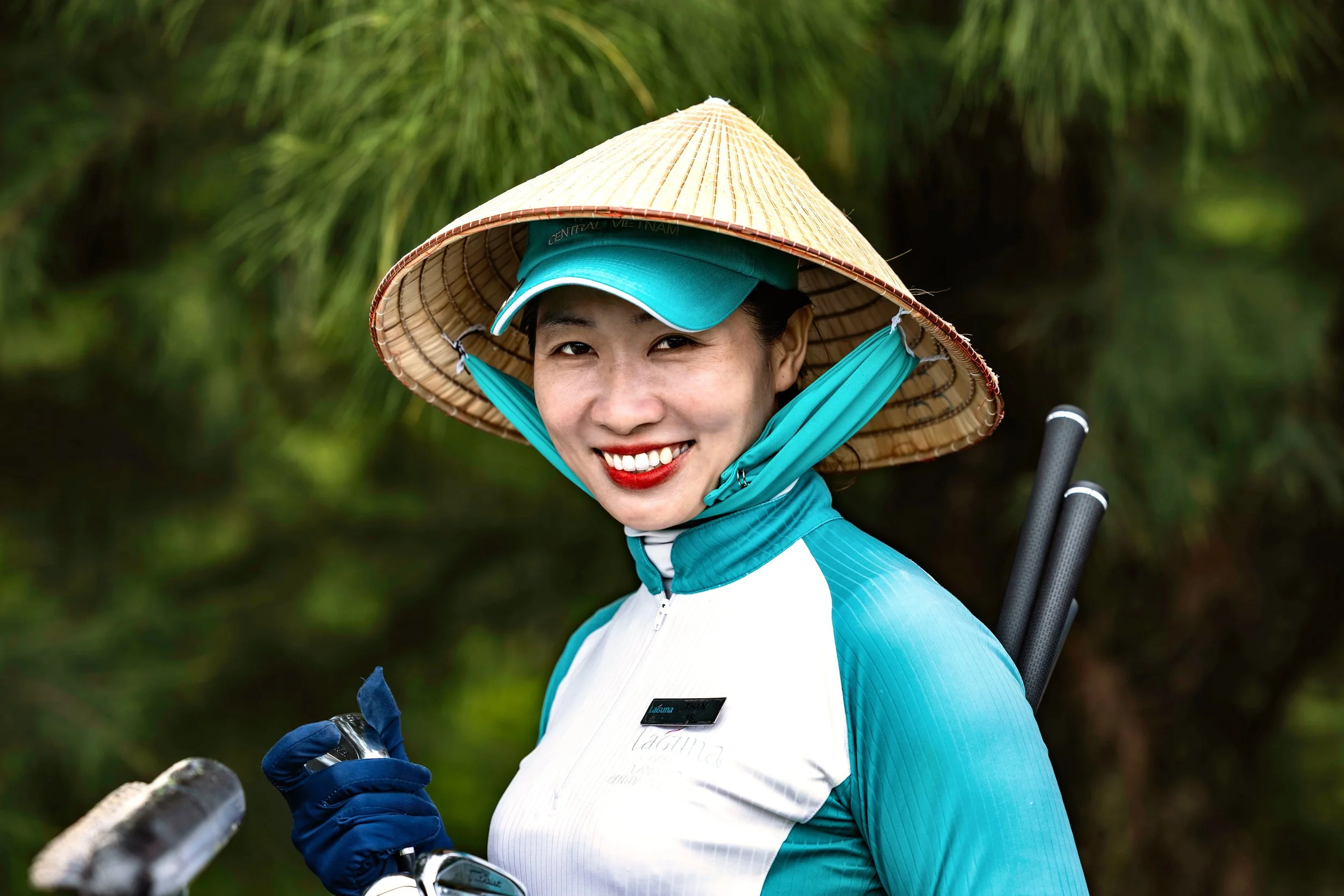Smiling woman wearing a traditional Vietnamese conical hat, a teal cap, a blue and white athletic jacket, and blue gloves, holding a golf club outdoors with green foliage in the background.