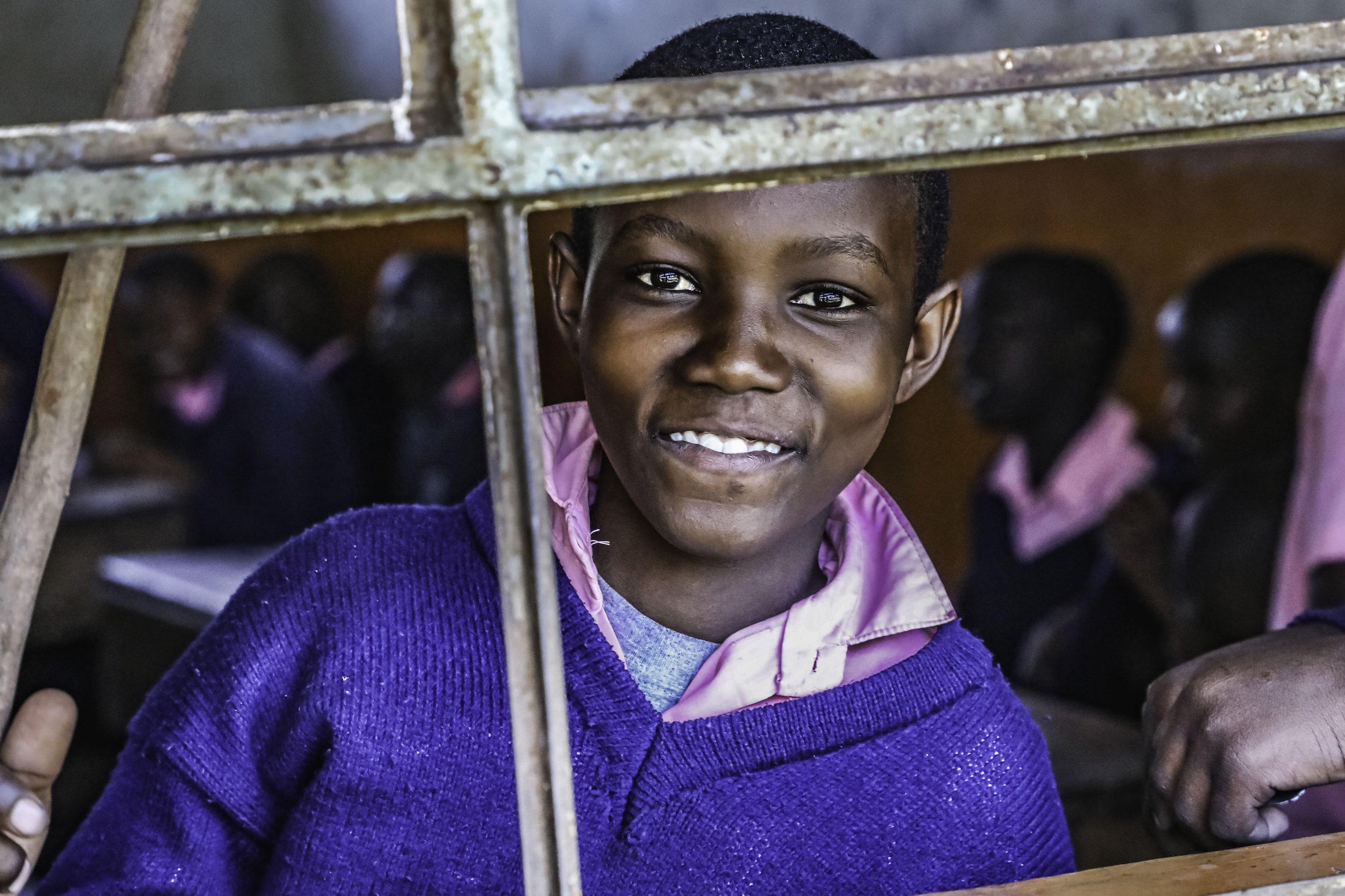 A young boy with short hair smiling and looking through a rusty metal fence, with blurred children in the background.