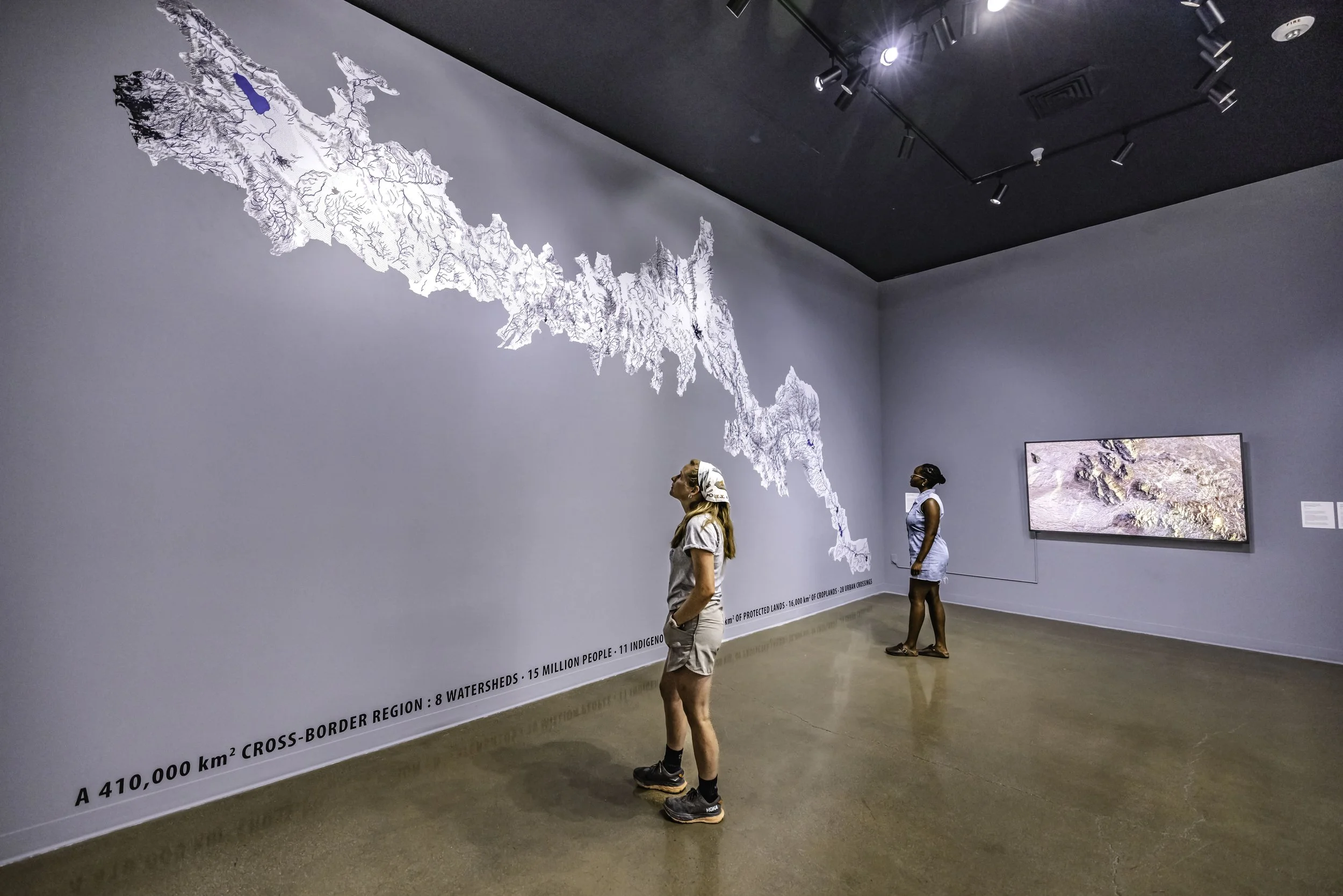 Three women are observing a large map of a region on a museum or gallery wall. The map depicts waterways, lakes, and terrain details. There is text at the bottom of the wall mentioning protected lands, indigenous lands, and watershed numbers.