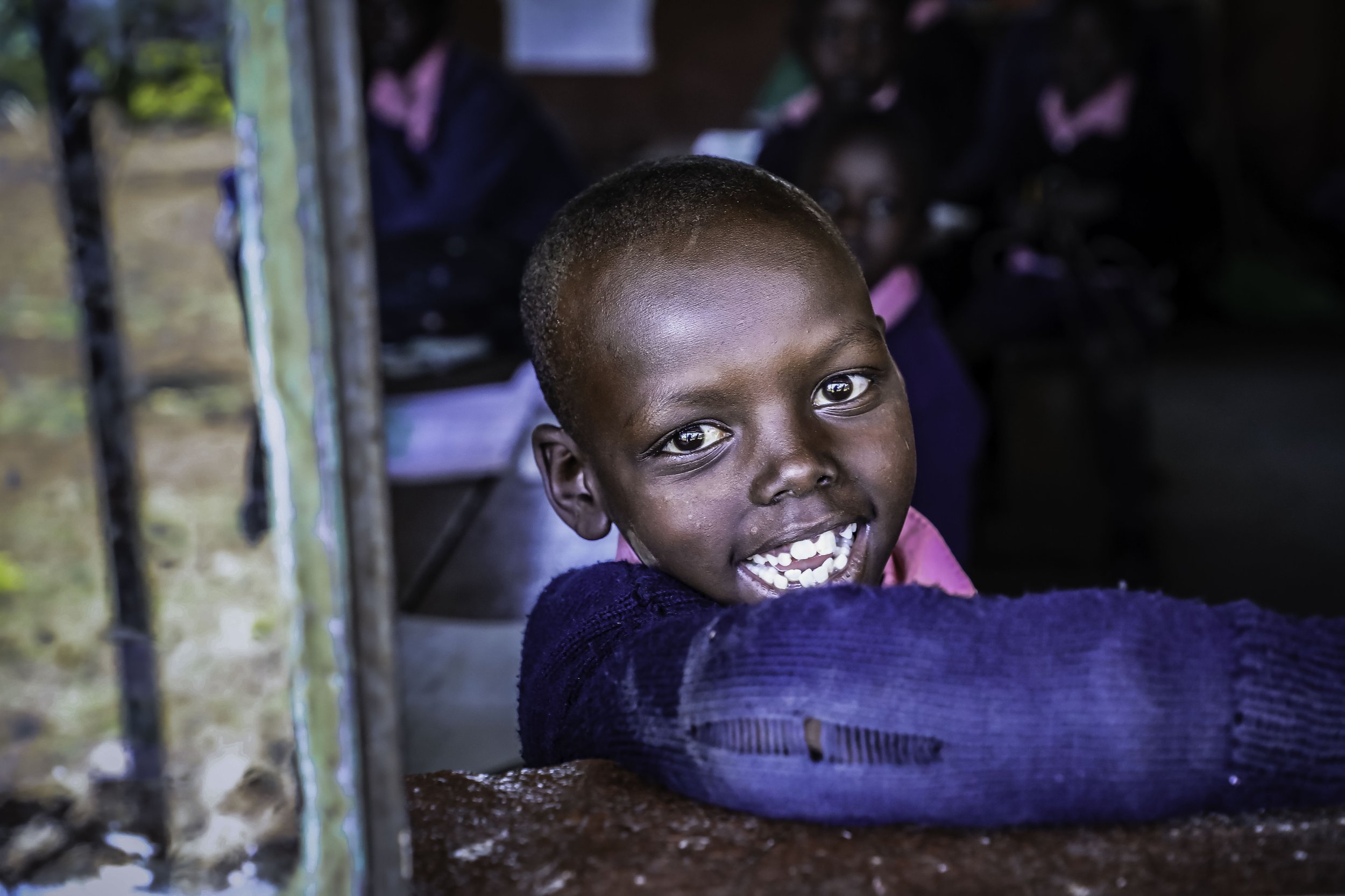 A young boy with a big smile, looking directly at the camera, inside a rustic wooden structure with other children in the background.