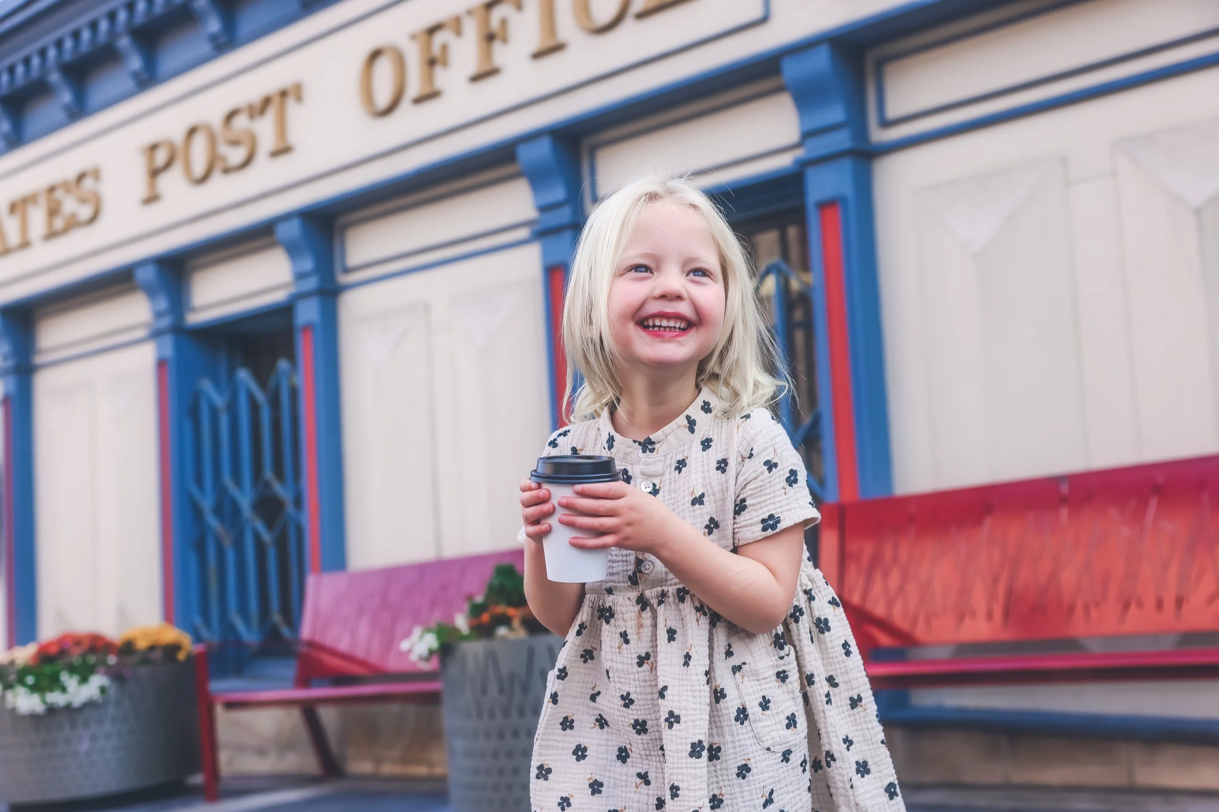A young girl with blonde hair smiling and holding a coffee cup standing in front of a colorful building with a sign that reads 'Post Office'.