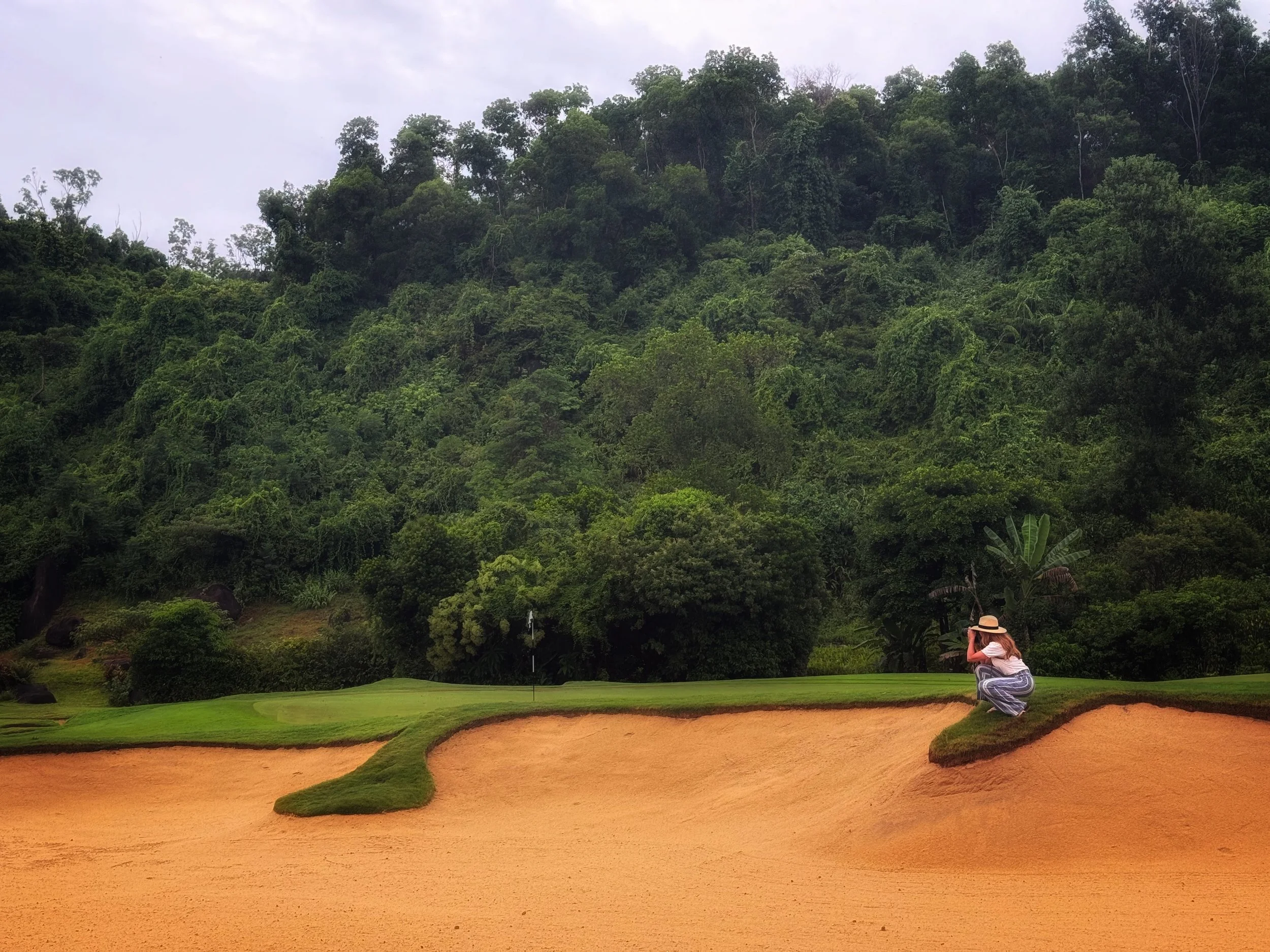 A woman in a hat taking a photo of the golf course with a view of dense green forest and cloudy sky in the background.