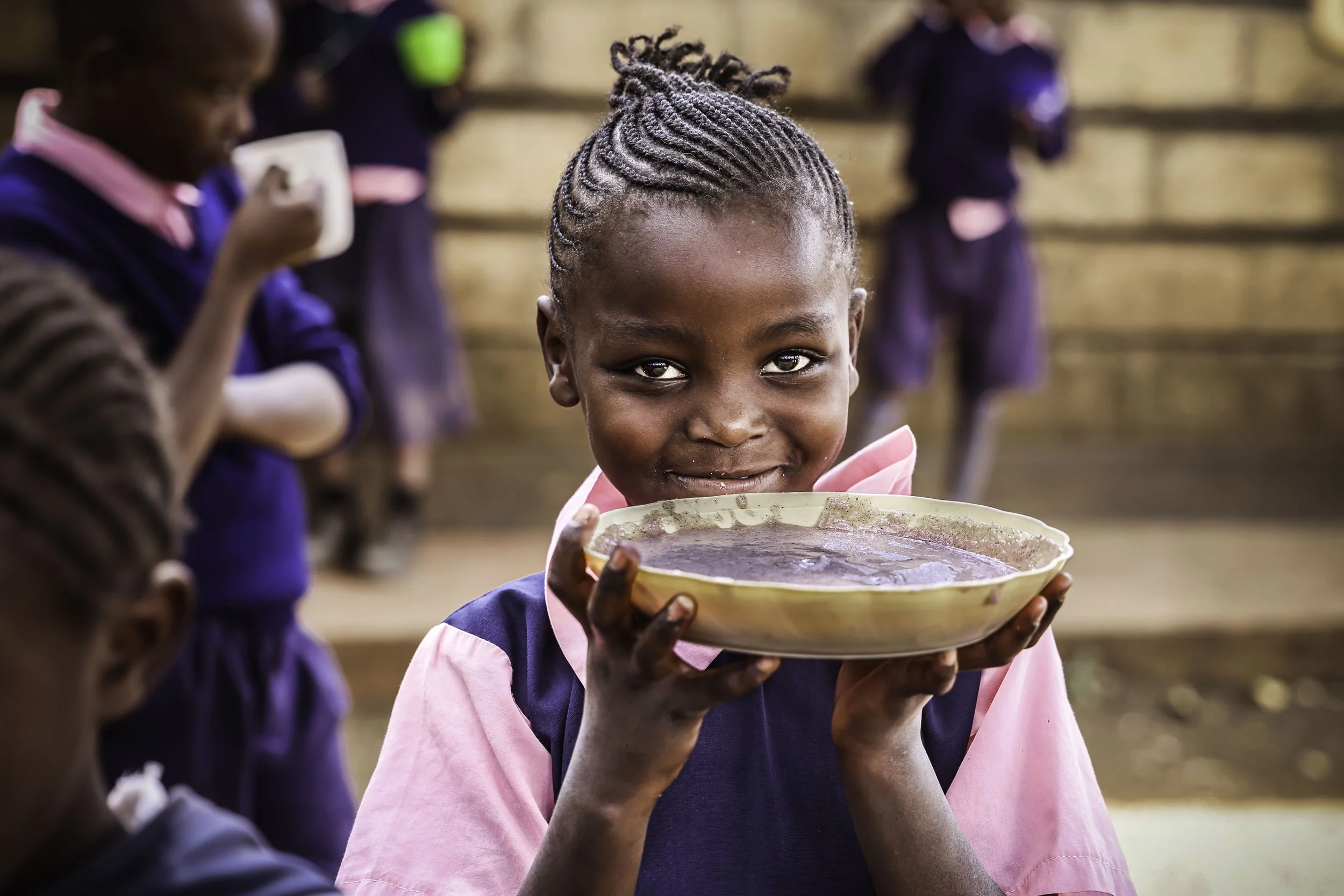 A young girl with braided hair holding a bowl of soup or porridge, smiling, with other children in the background in a school setting.