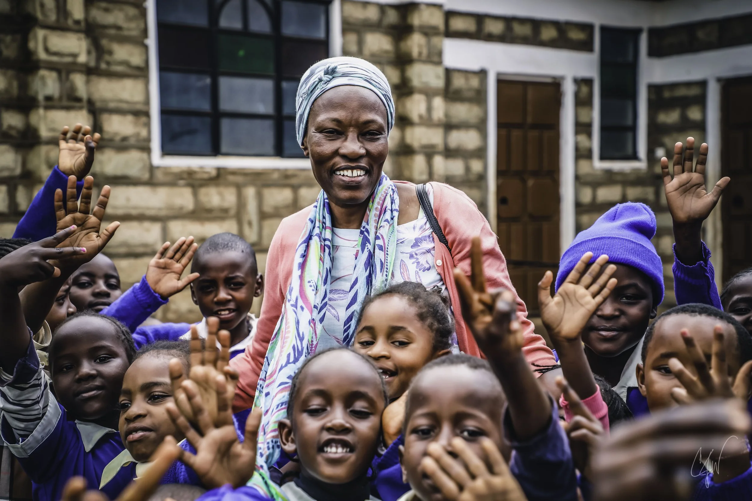 A woman wearing a headscarf and colorful clothing surrounded by smiling children, outside in front of a brick building with windows.