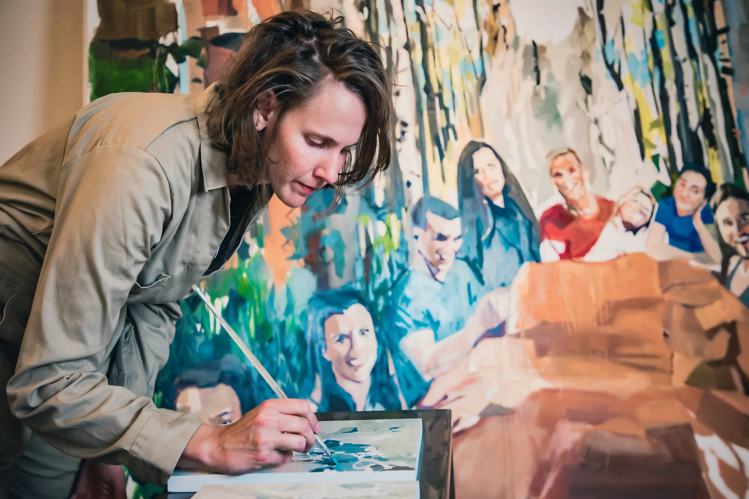 A woman with shoulder-length curly brown hair painting a group portrait on a large canvas. The background features a colorful mural with several faces of diverse individuals.