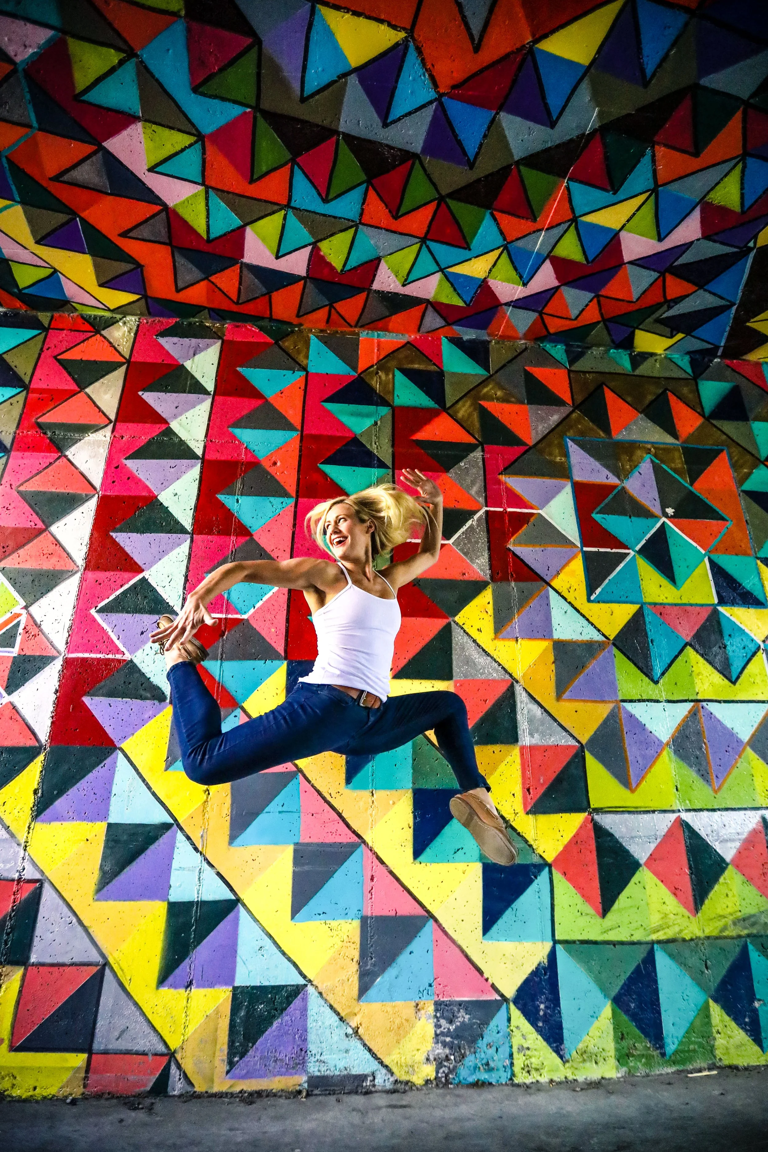 A woman with blonde hair in a white tank top and blue jeans performing a jump in front of a colorful geometric painted wall.