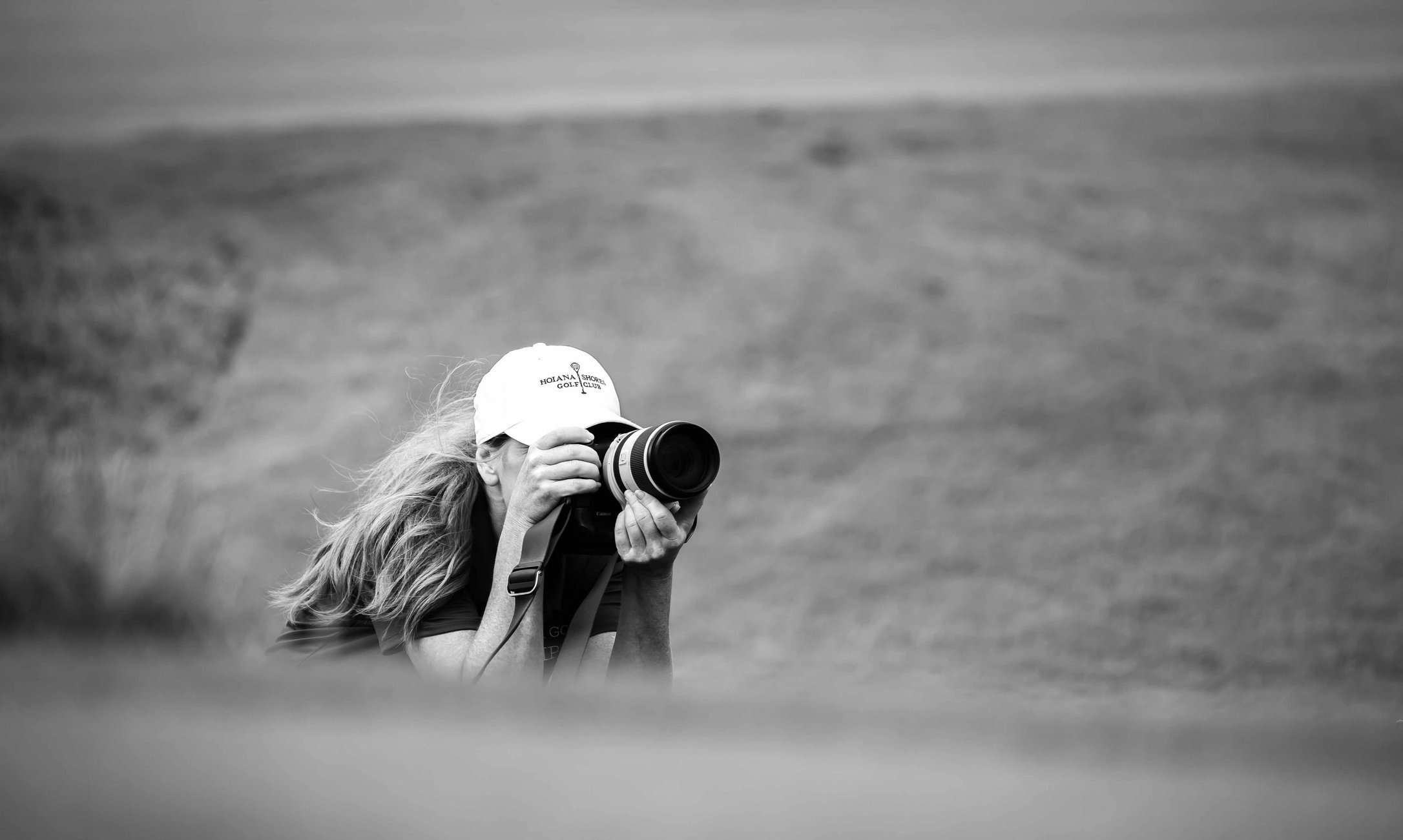 A woman with long hair, wearing a baseball cap that says 'Holana Golf Shore Club,' is taking a photograph with a professional camera outdoors on a grassy field.