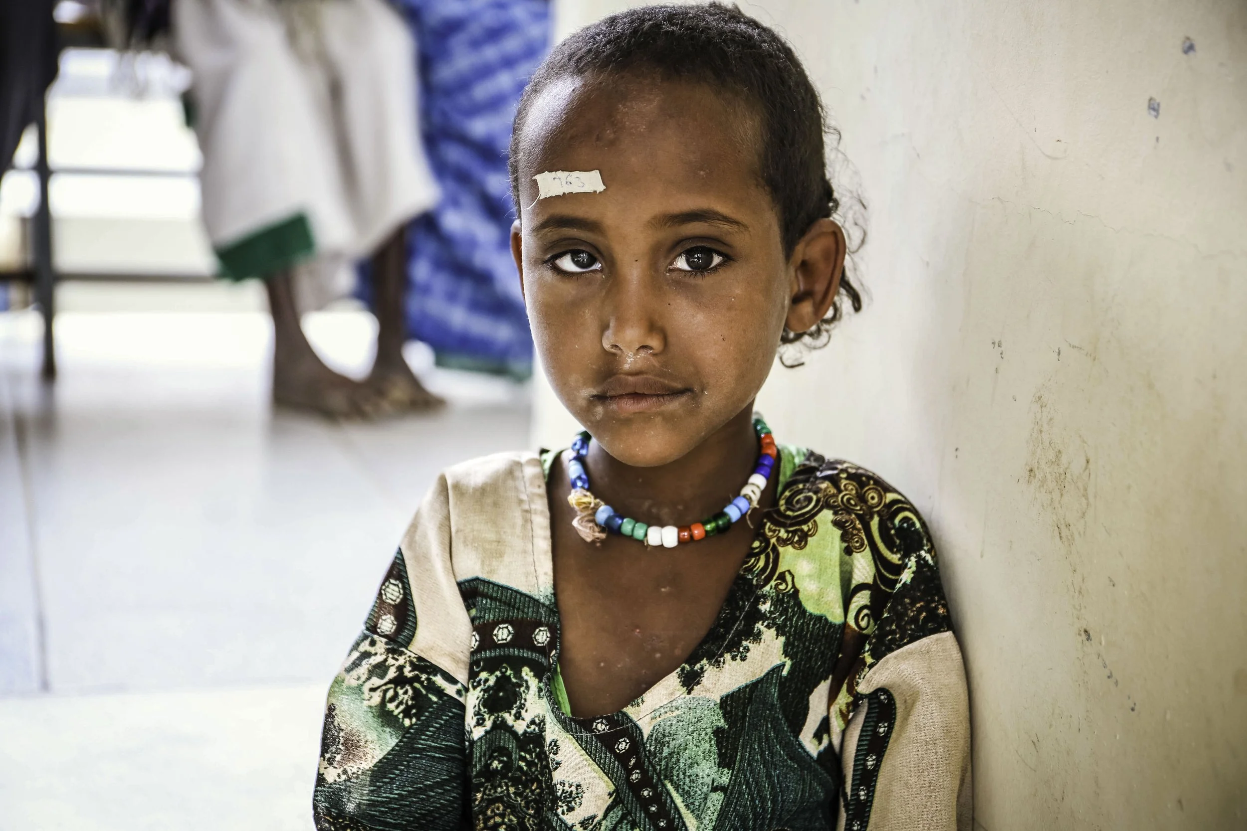 A young girl with dark curly hair and brown skin sitting against a wall in what appears to be a school or community center. She is wearing colorful beaded necklaces and a patterned dress with green, black, and beige colors. There is a bandage on her 