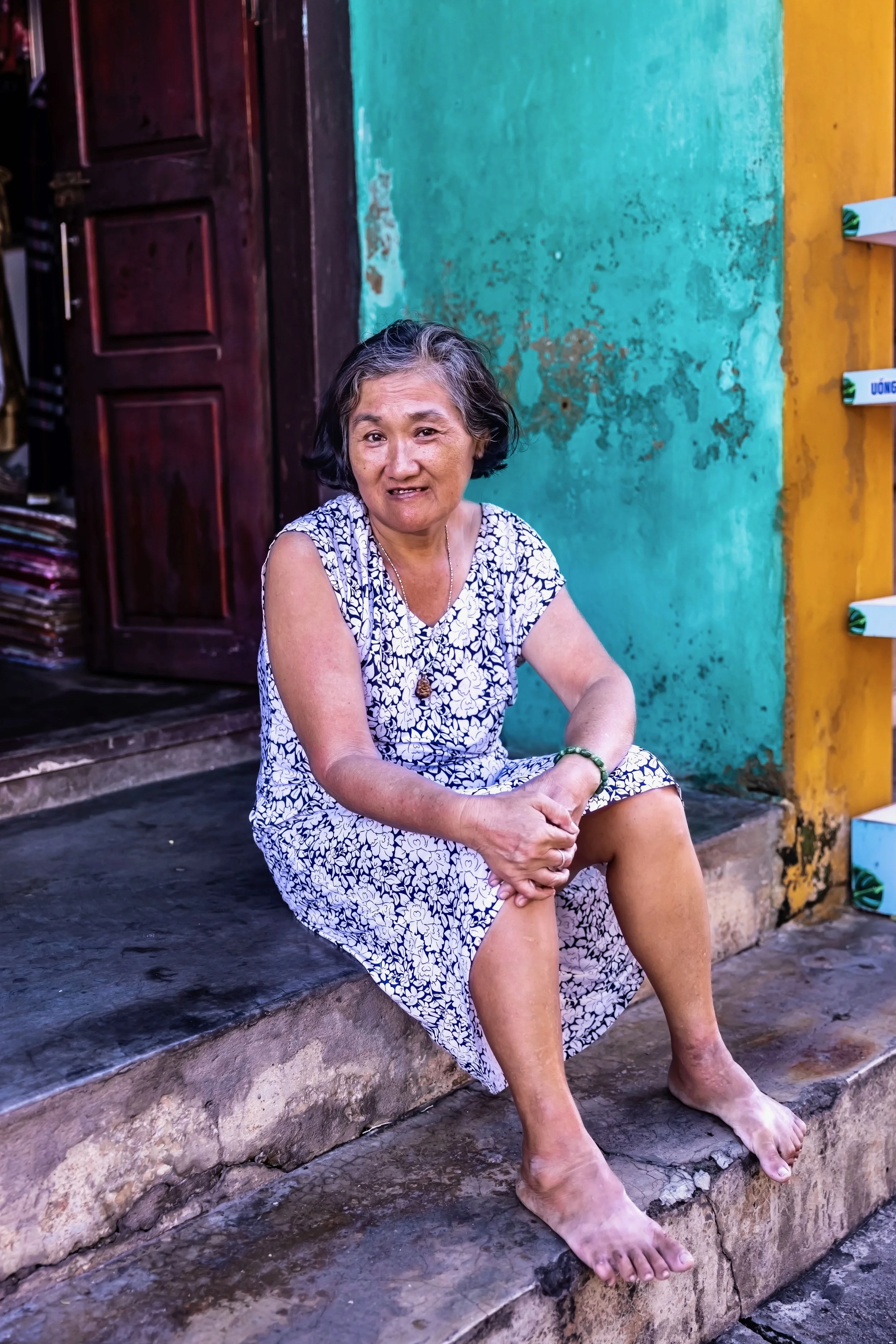 An elderly woman with short gray hair sitting on stone steps with her legs crossed, wearing a sleeveless floral dress, barefoot, and wearing a green bracelet, with peeling turquoise and yellow walls and a wooden door in the background.