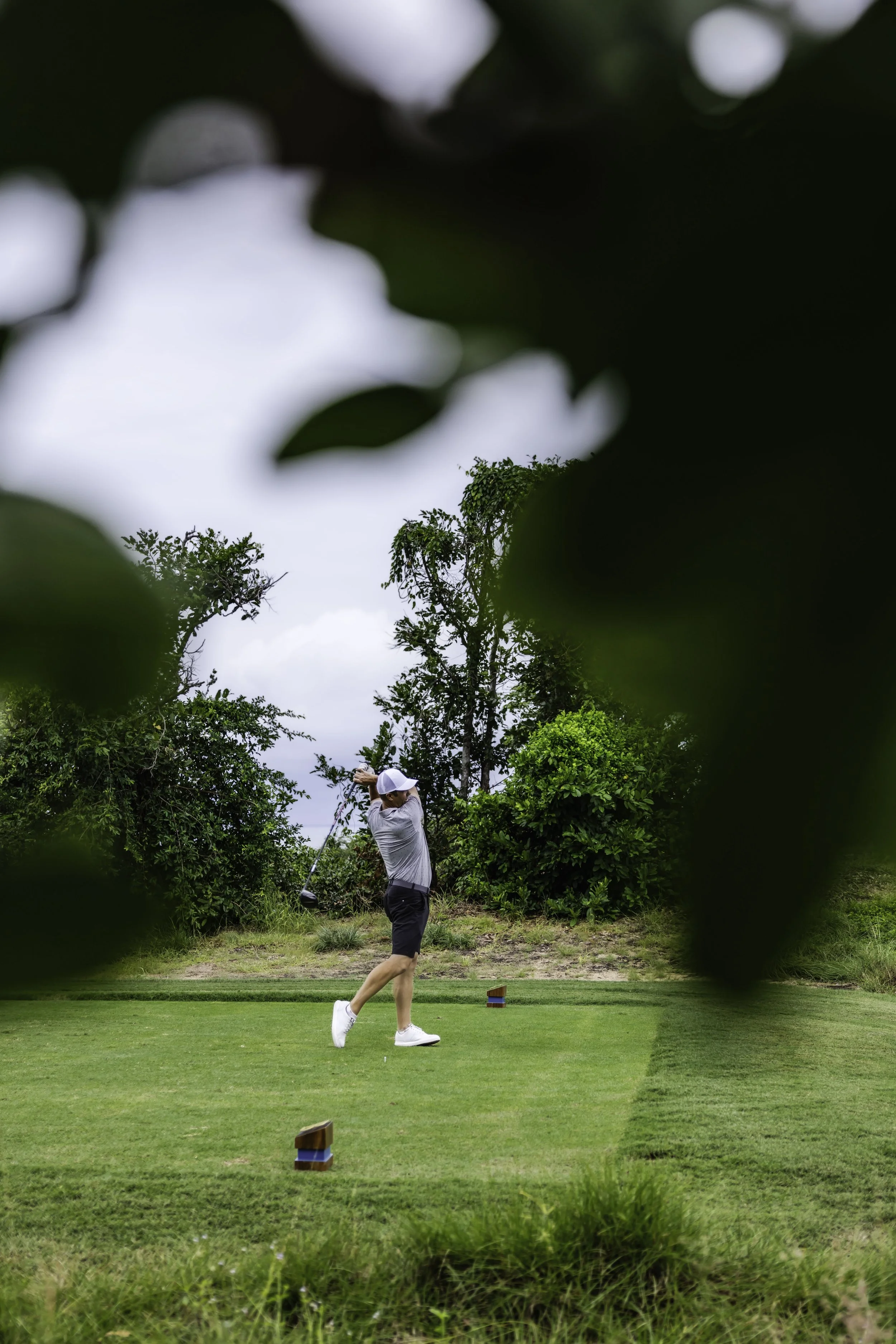A person playing golf on a lush green course, swinging a golf club. The scene is surrounded by trees and greenery, with partly cloudy skies above.