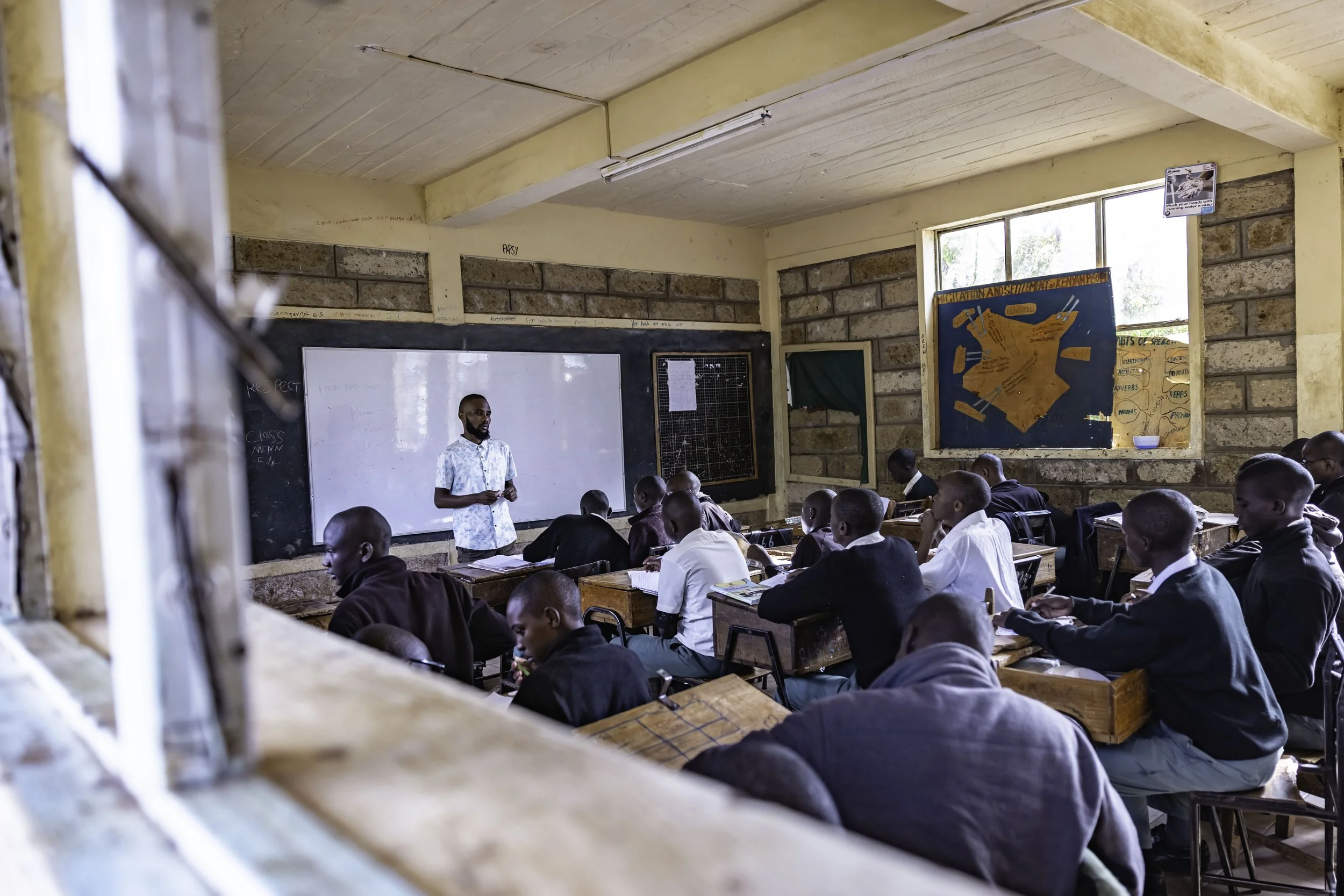 A classroom with students seated at desks, facing a teacher standing near a whiteboard, with educational posters on the walls and a window on the side.