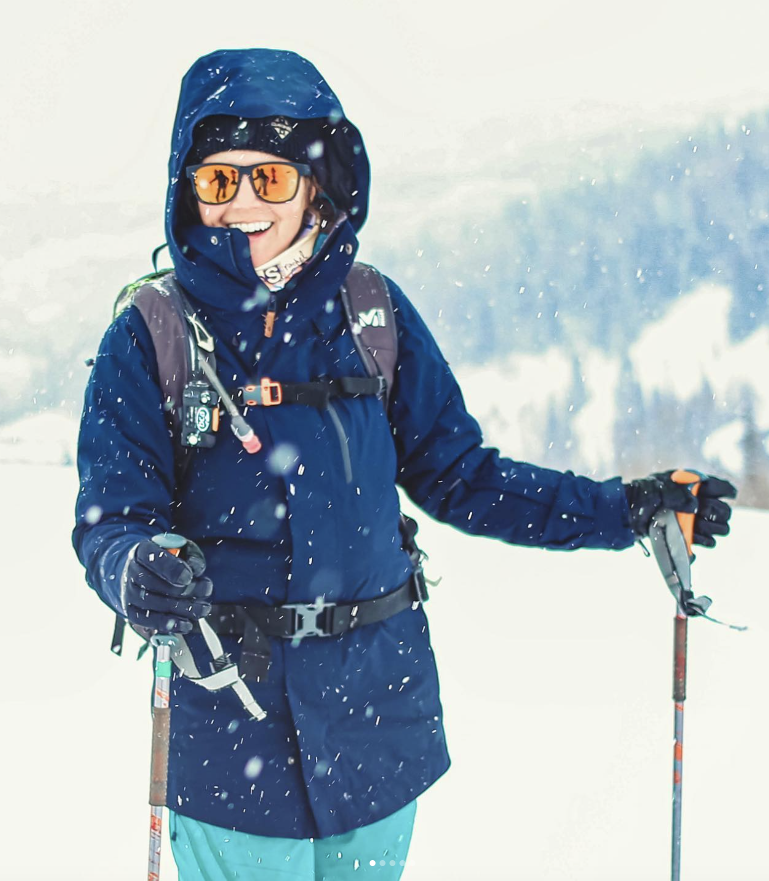Person in blue winter gear smiling on snowy mountain, holding ski pole, wearing reflective sunglasses, snow falling around.