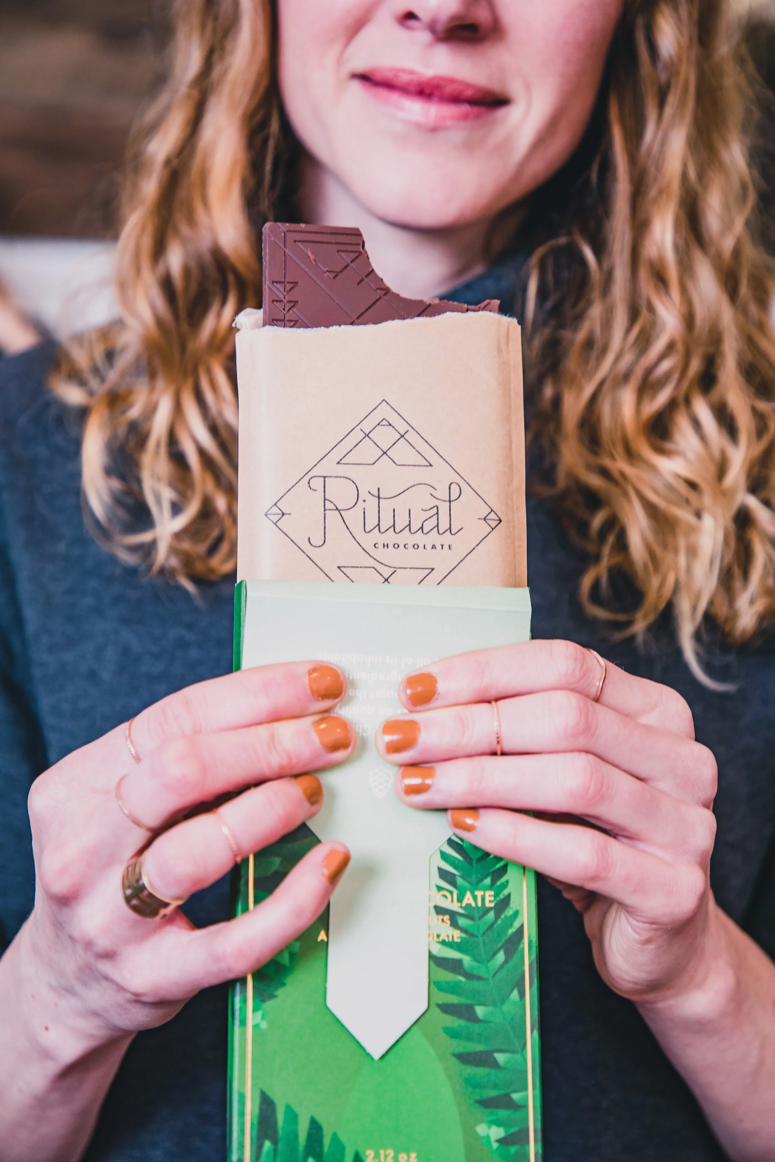 A woman with curly red hair holding a chocolate bar and a green and white packaged product in front of her chest. The chocolate bar is partially unwrapped, revealing a piece of chocolate. The woman's face is partially visible, with her lips slightly 