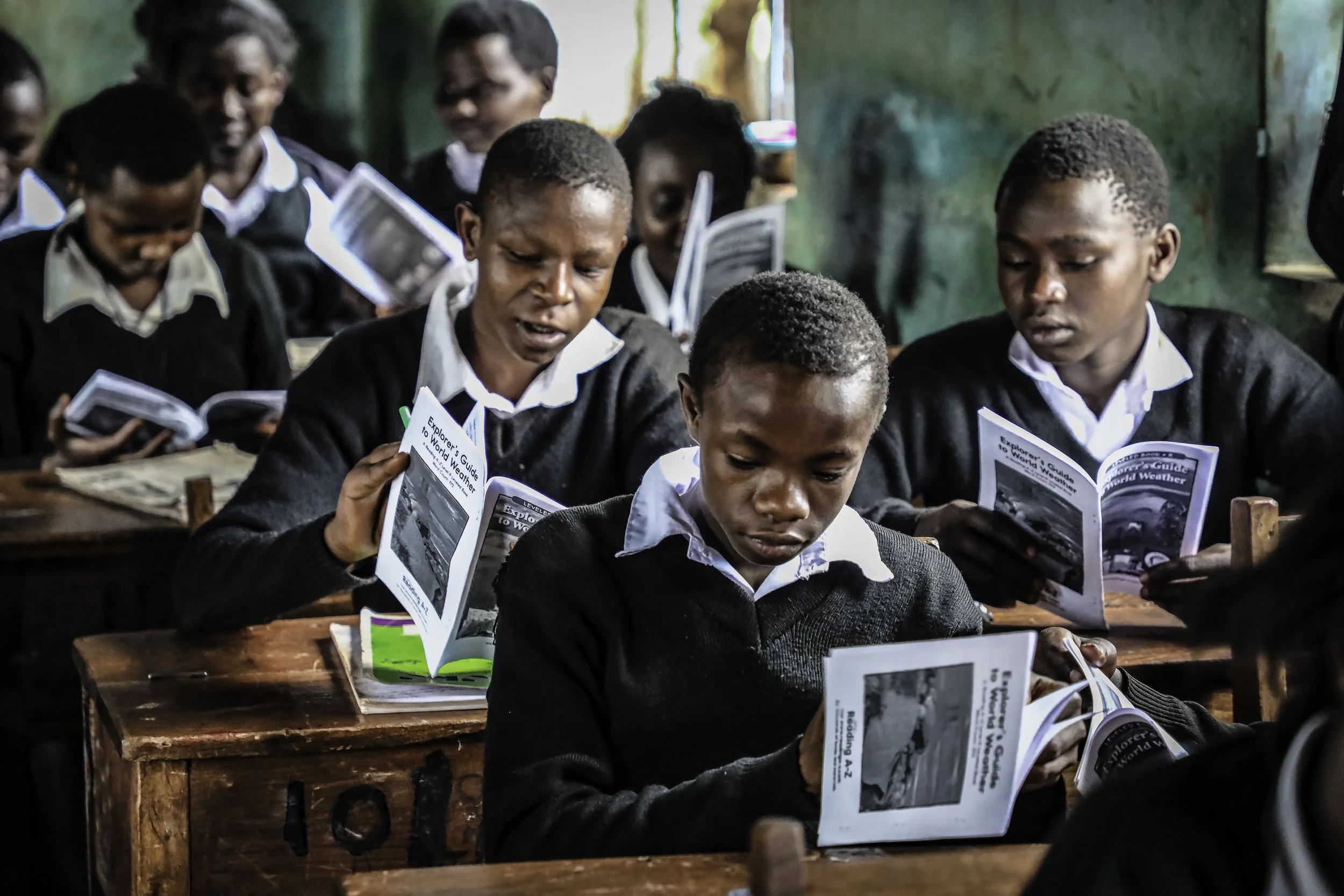 A group of young students in uniform reading books in a classroom with green walls.