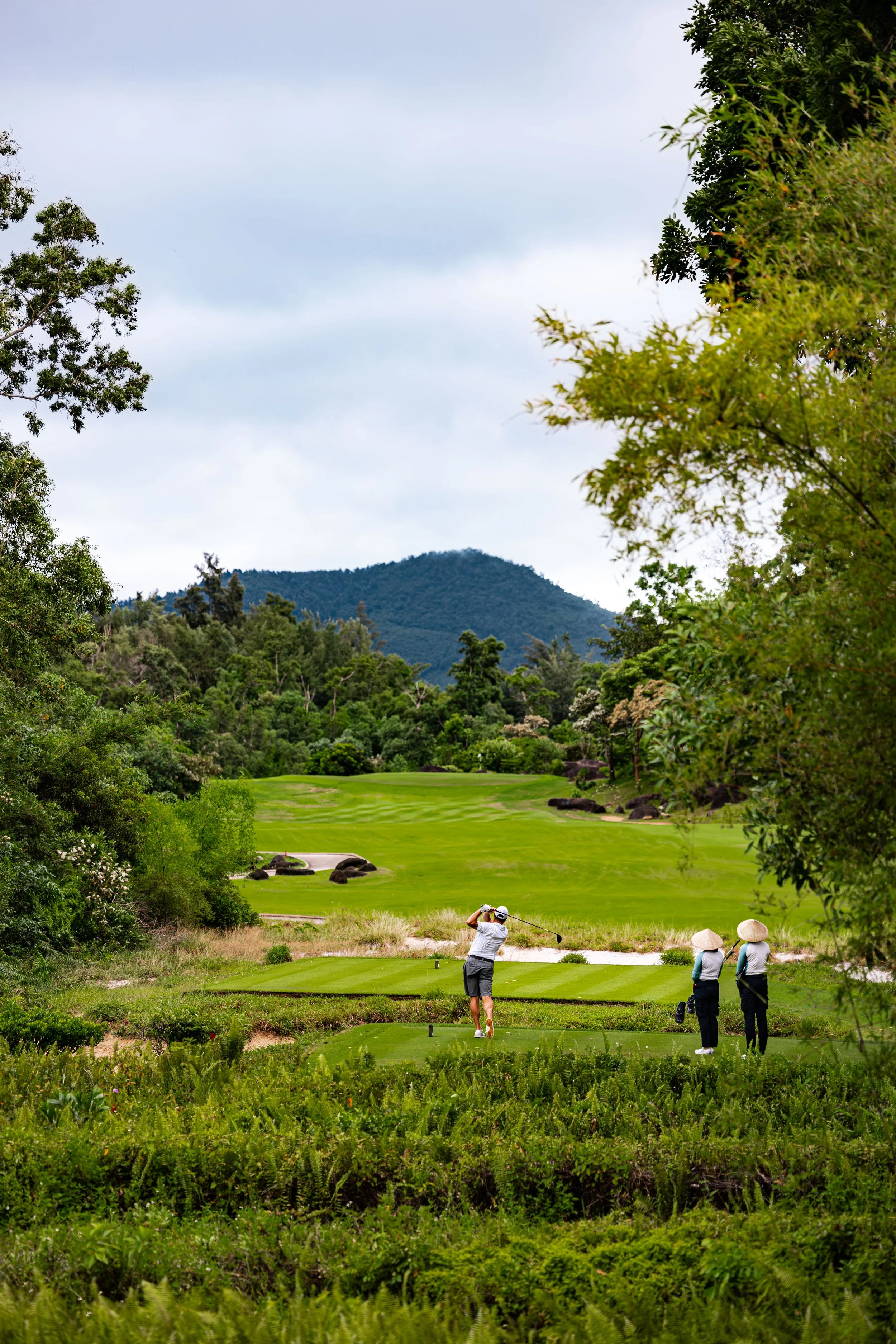 A golfer swings a club on a lush golf course with two spectators wearing traditional Asian straw hats watching nearby, surrounded by green trees and distant mountains under a cloudy sky.