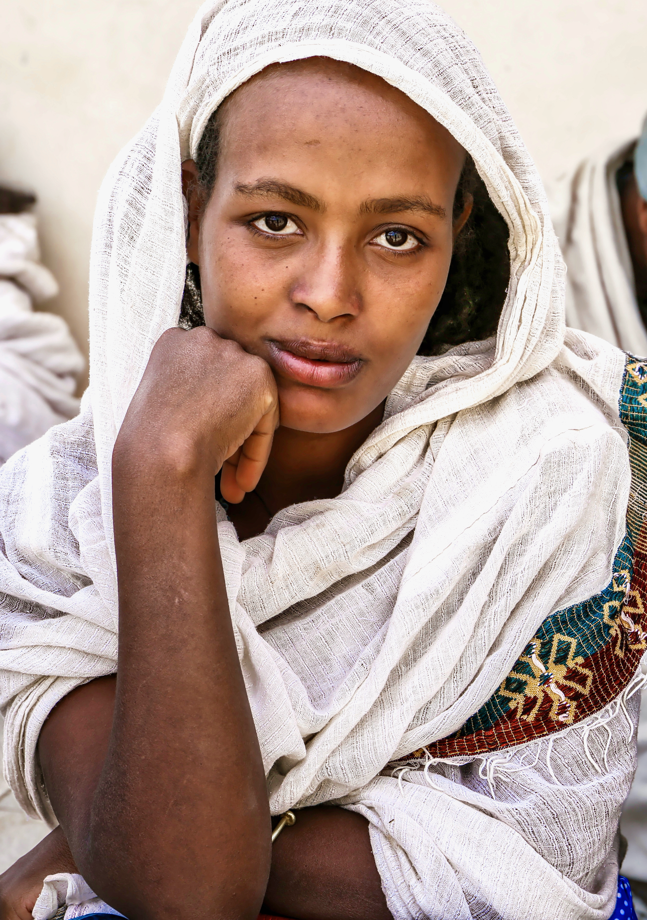 Close-up of a woman with medium brown skin, wearing a white headscarf and wrap, sitting with her face resting on her hand, looking directly at the camera.