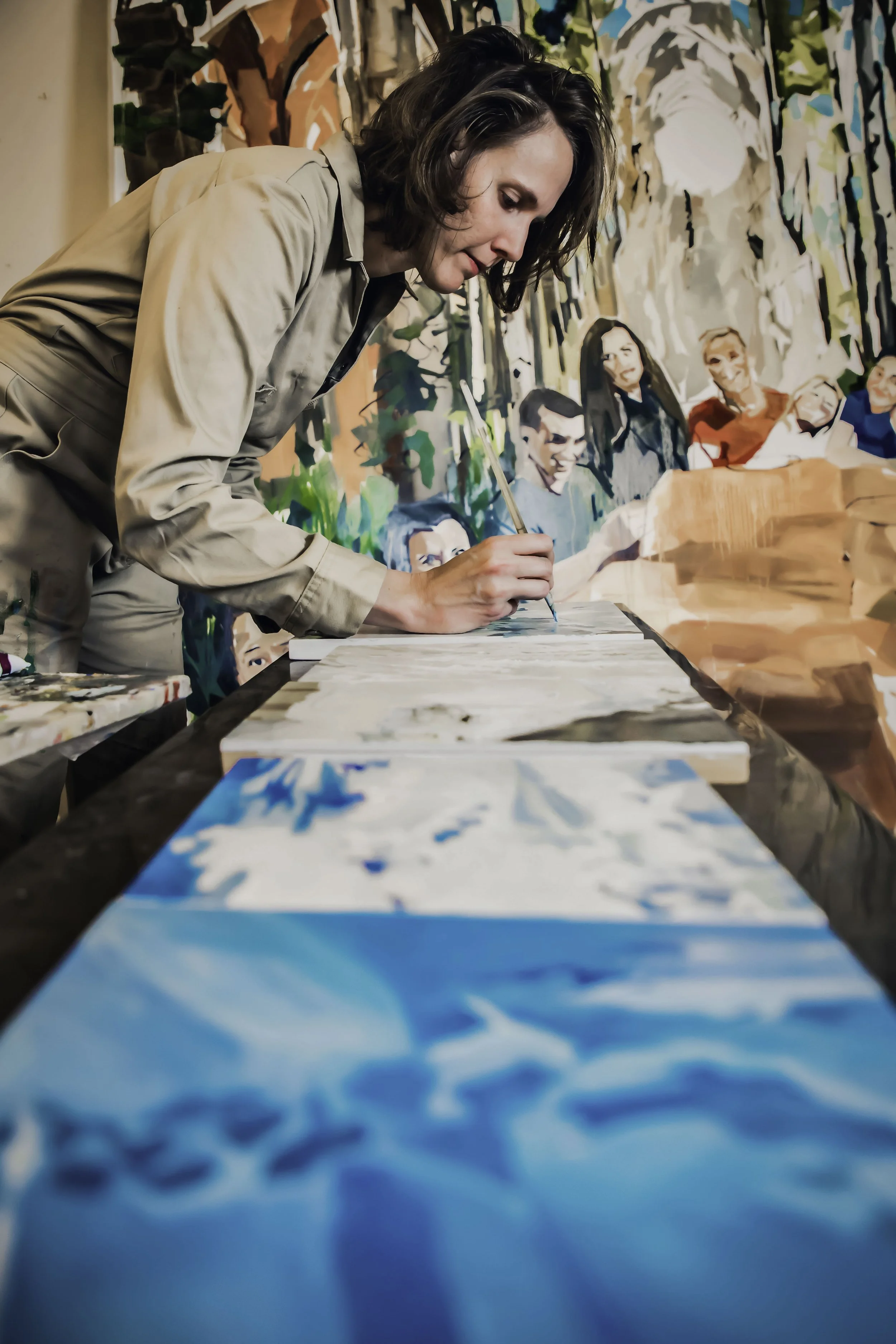 A woman with shoulder-length brown hair, wearing a beige shirt, is leaning over a table painting on a wood surface, with a colorful mural painted on the wall behind her.