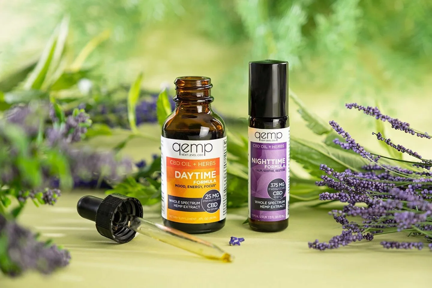 Commercial product photography for Qemp Day and Night CBD tinctures. Two dropper bottles are styled with fresh ferns and purple lavender blooms under dramatic, high-contrast studio lighting for a wellness brand.