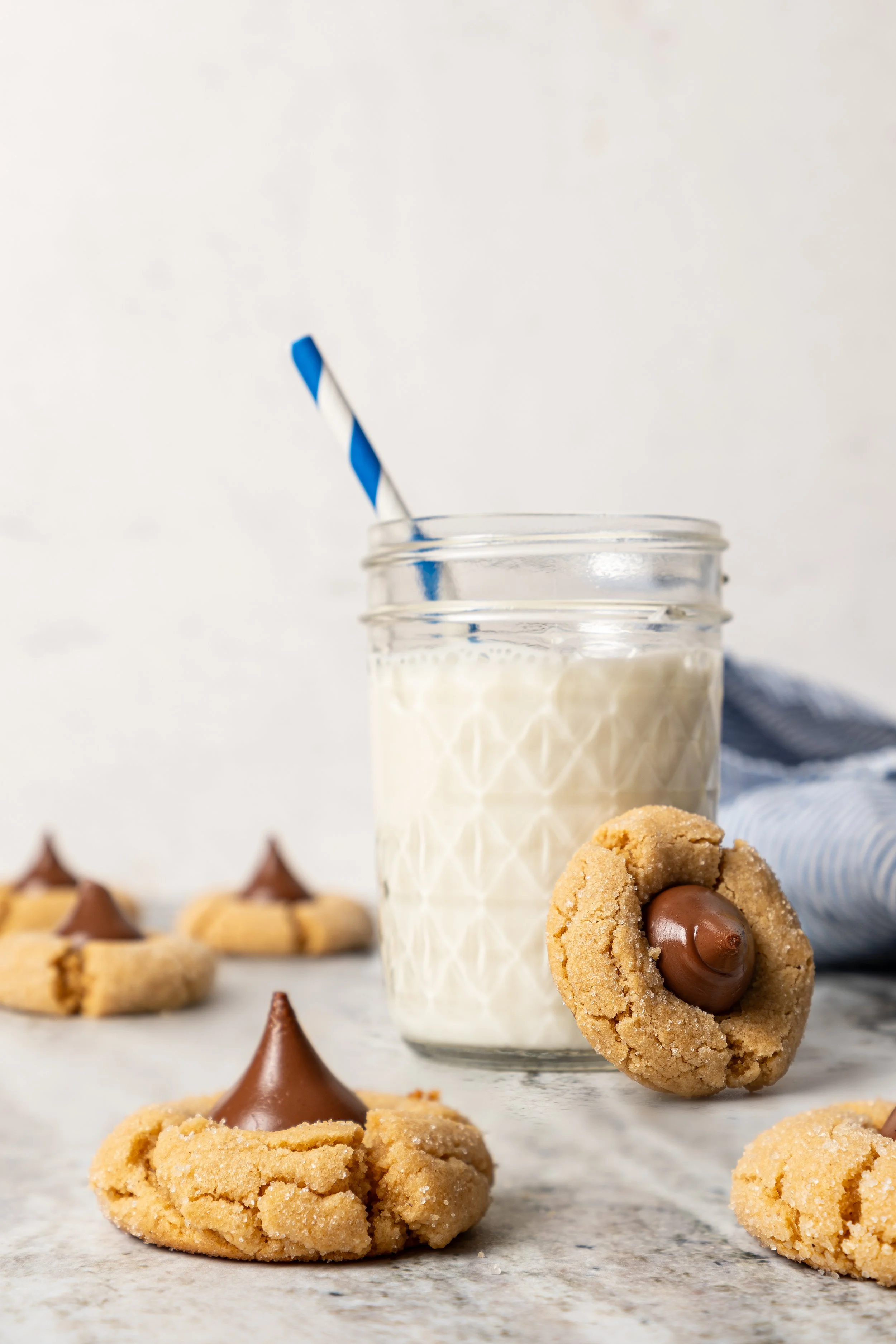 Peanut butter blossom cookies from a Pennsylvania Grange Cookbook recipe on the JodiLoves blog. A sugar-dusted cookie with a chocolate kiss candy center sits on marble next to a vintage glass of milk with a striped straw.
