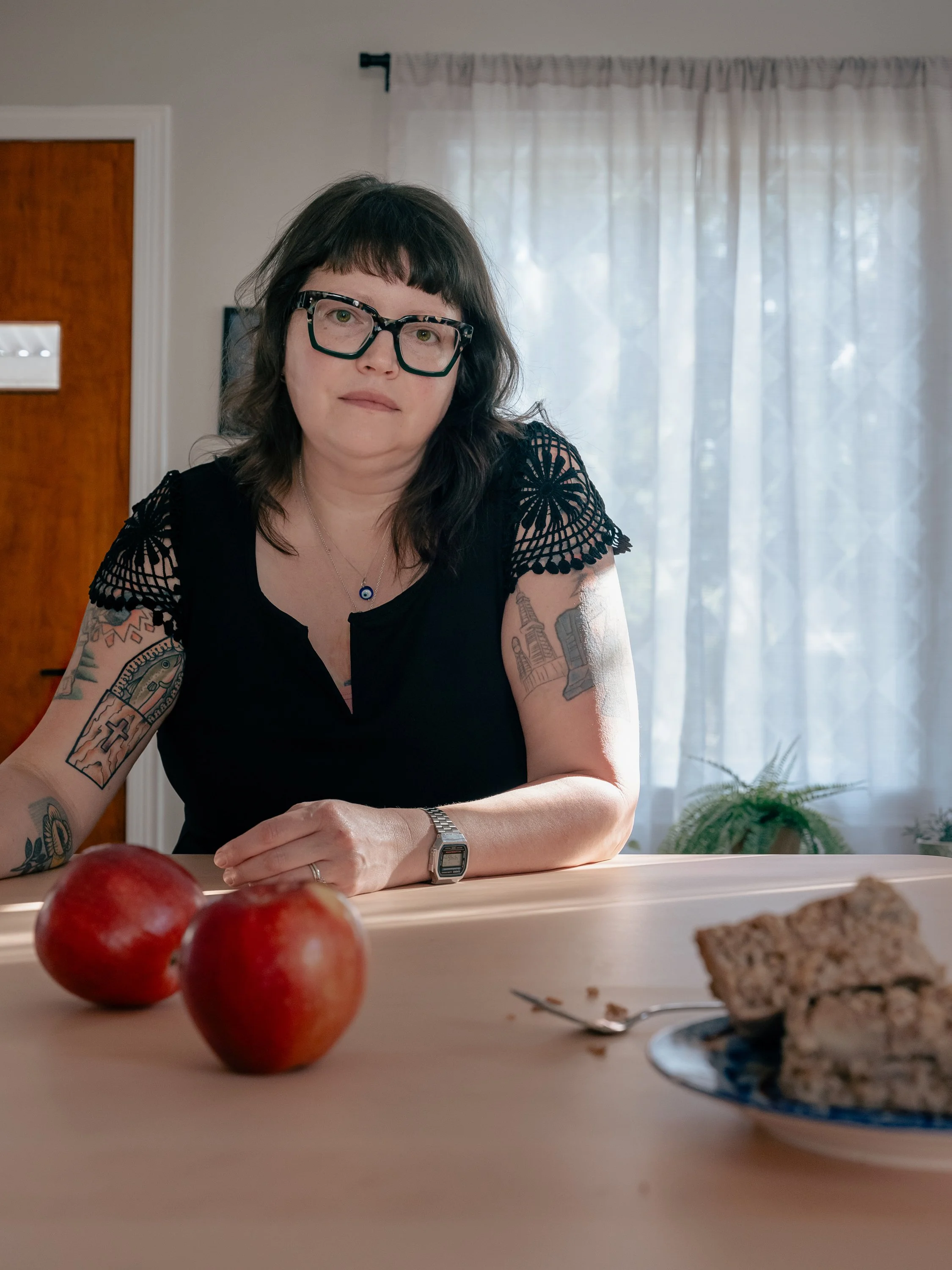 Jodi Boatman, professional food photographer and commercial digital retoucher at Brilliant Pixel Imaging. Portrait of the artist sitting at a wooden studio table styled with fresh red apples and baked goods under soft, natural window light