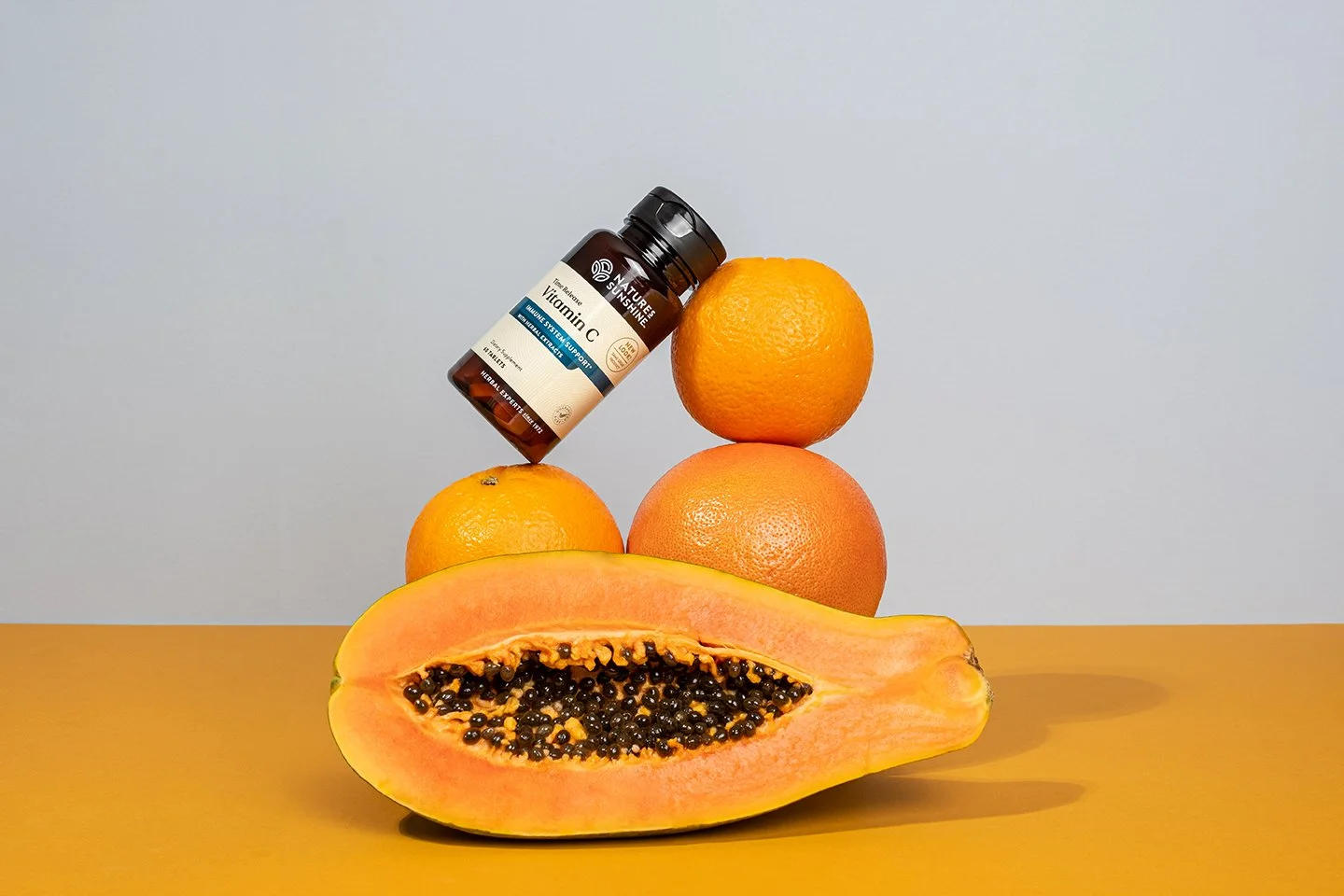 Product shot showcasing a conceptual pyramid stack of fresh citrus fruit balancing a vitamin supplement bottle. Studio lighting emphasizes textured orange peels and a halved papaya against a clean dual-tone backdrop.