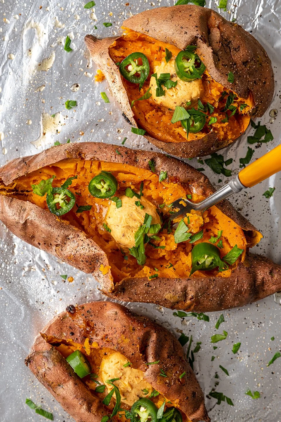 Macro food photography of seasoned baked sweet potatoes sliced on foil. Features a melting scoop of gourmet compound butter, fresh chopped parsley, and jalapeño slices with a rustic yellow-handled fork for a culinary campaign.