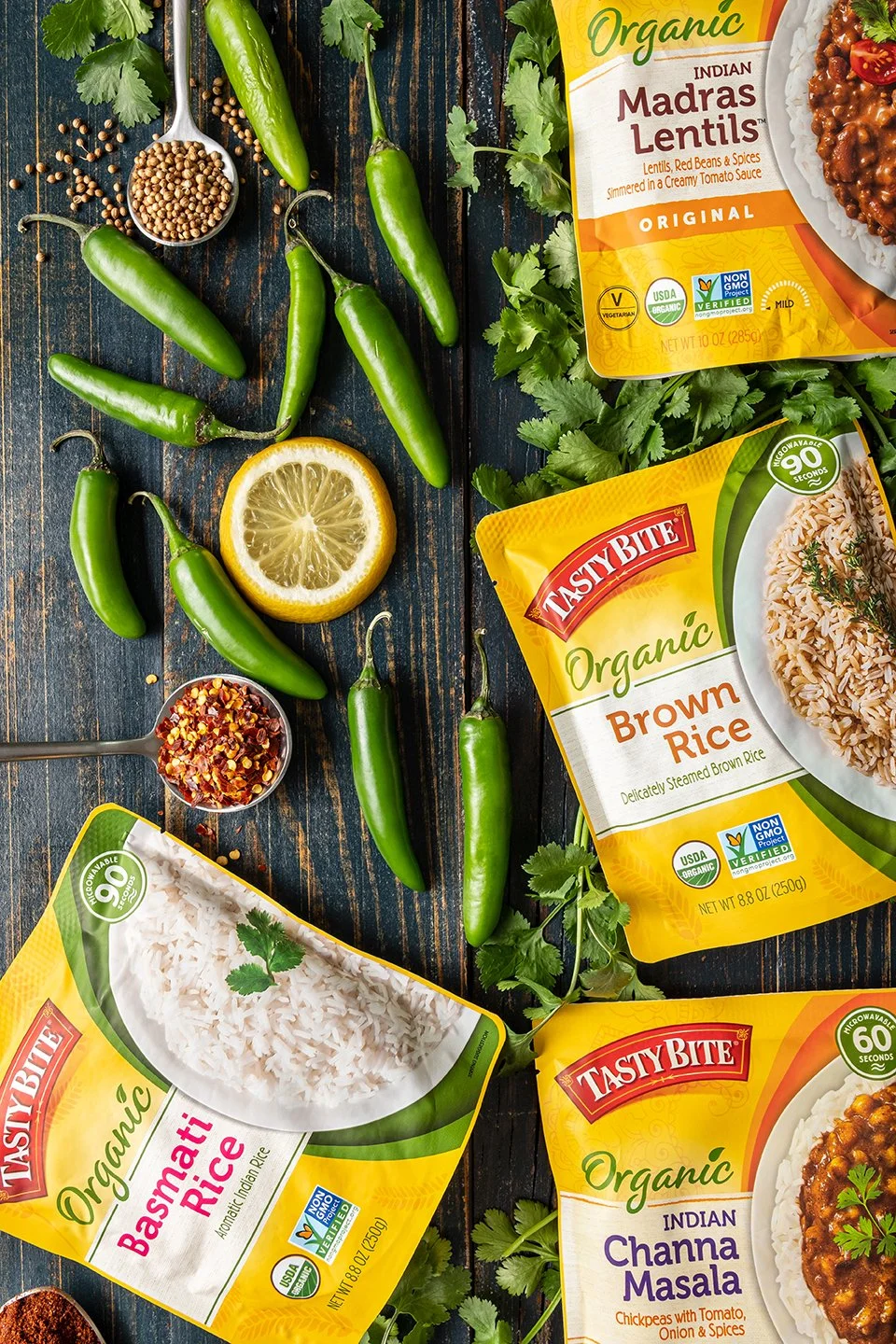 High-end CPG food styling featuring organic Indian meal pouches. Overhead studio lighting highlights glossy fresh green chilis, red pepper flakes, and vibrant herbs for a balanced, high-contrast recipe composition.
