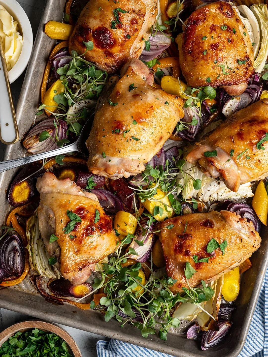 Overhead commercial food photography of a sheet pan meal featuring crispy roasted chicken thighs, red onions, carrots, and cabbage garnished with microgreens, styled with a small side bowl of premium butter.