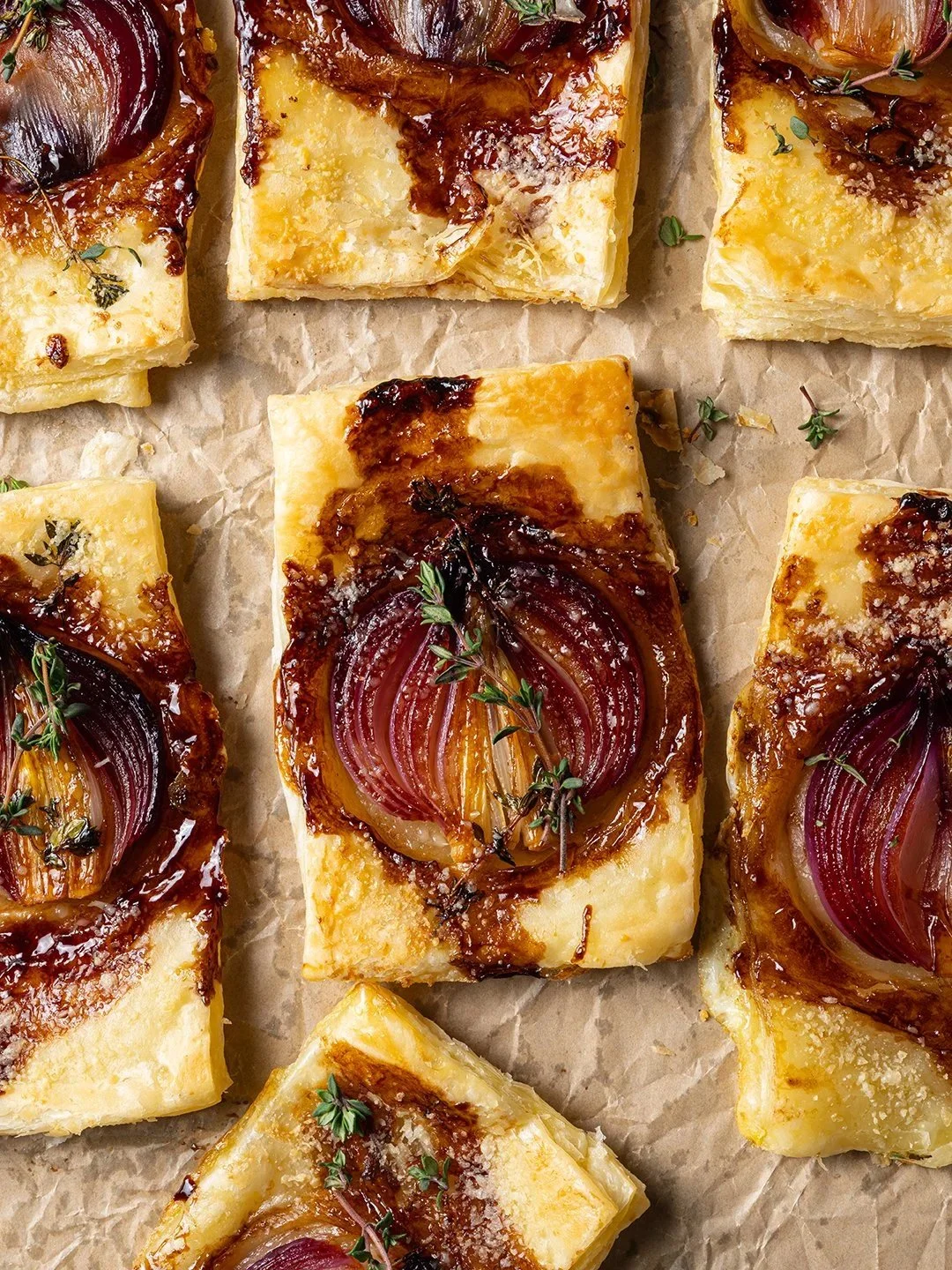 Top-down food styling of savory puff pastry tarts topped with caramelized red onion halves and fresh thyme sprigs, arranged symmetrically on crumpled brown baking parchment paper for a culinary campaign.