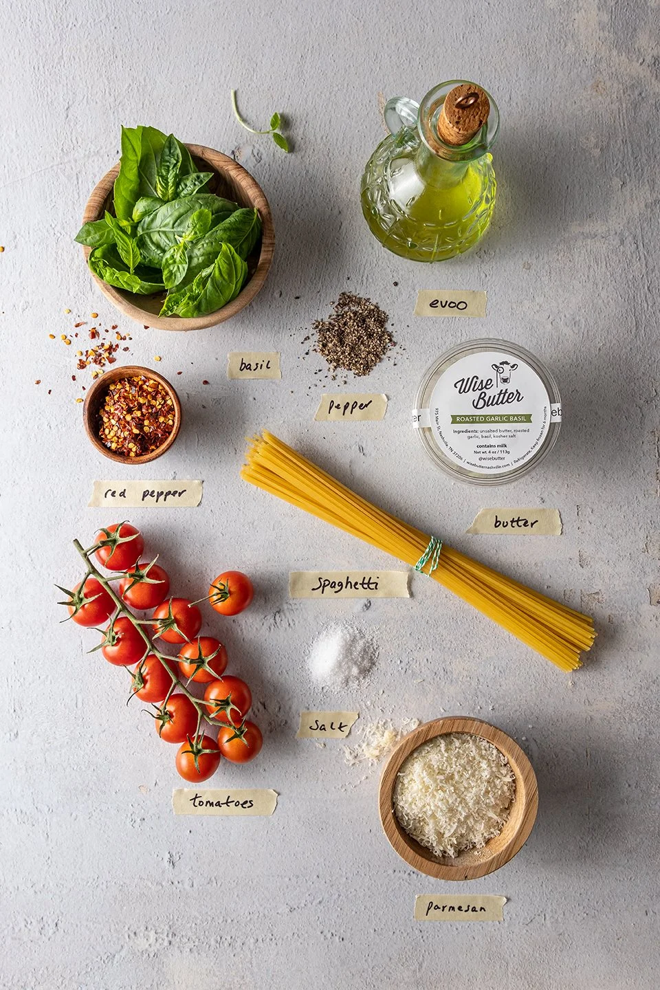 Overhead food styling layout for an Italian pasta dish campaign. Clean studio lighting highlights grated parmesan cheese, red pepper flakes, and salt arranged symmetrically with a tub of Wise Butter gourmet infused butter.