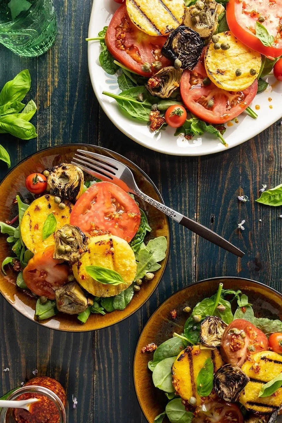 Editorial food photography of a Mediterranean summer salad flat lay with grilled polenta rounds, tomatoes, and marinated artichokes. A silver fork and chili oil jar accent the dark wood table.