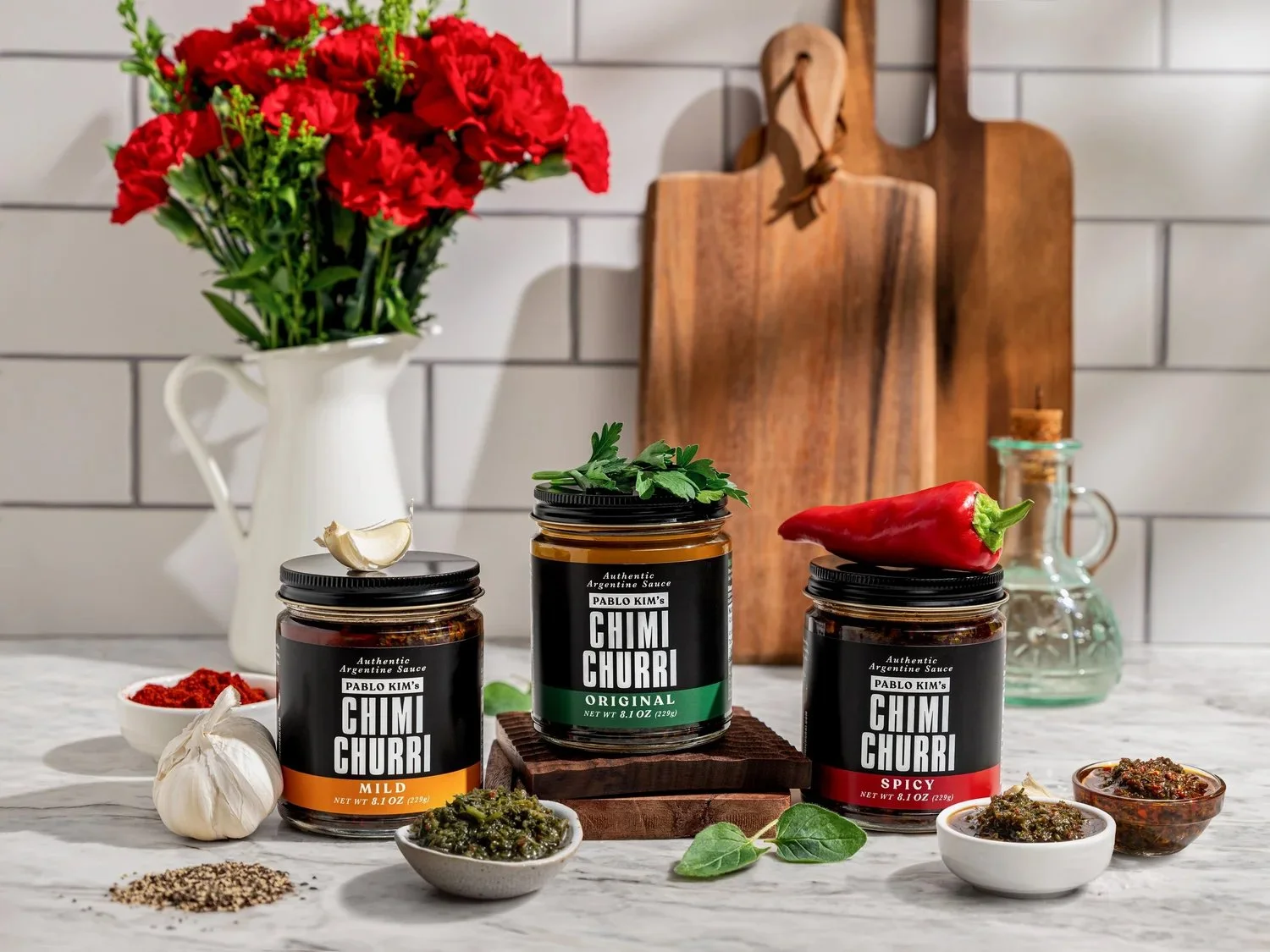 Commercial food product photography for Pablo Kim's Chimi Churri. Three jars of Argentine sauce (Mild, Original, Spicy) styled with garlic, fresh herbs, and a red pepper on a marble counter against white subway tile.